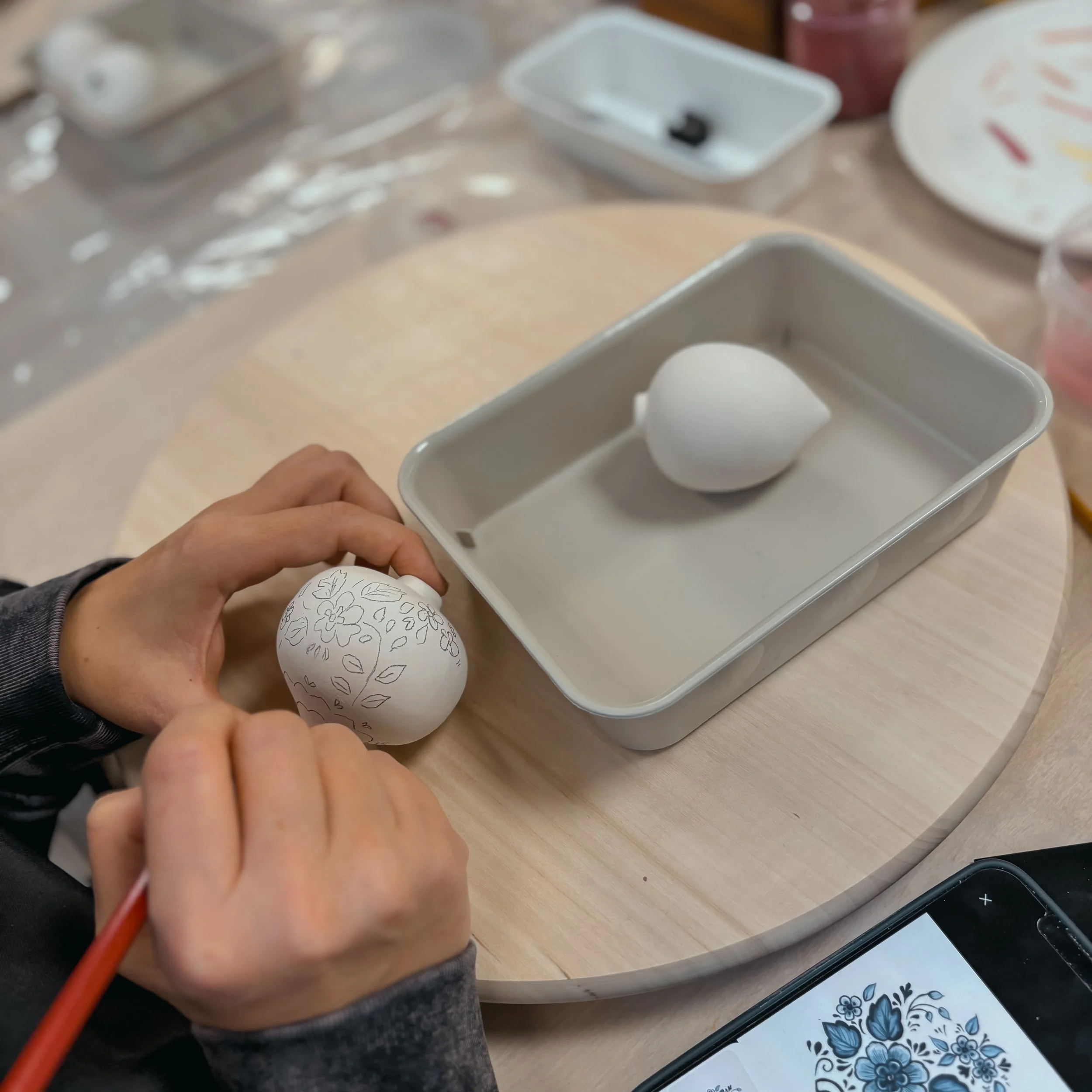 Una persona disegna un motivo floreale su un uovo di ceramica bianca con un pennarello nero, mentre un'altra parte dell'uovo è già decorata con un disegno floreale. L'uovo decorato è tenuto in mano, mentre l'altra parte dell'uovo si trova in una tegl
