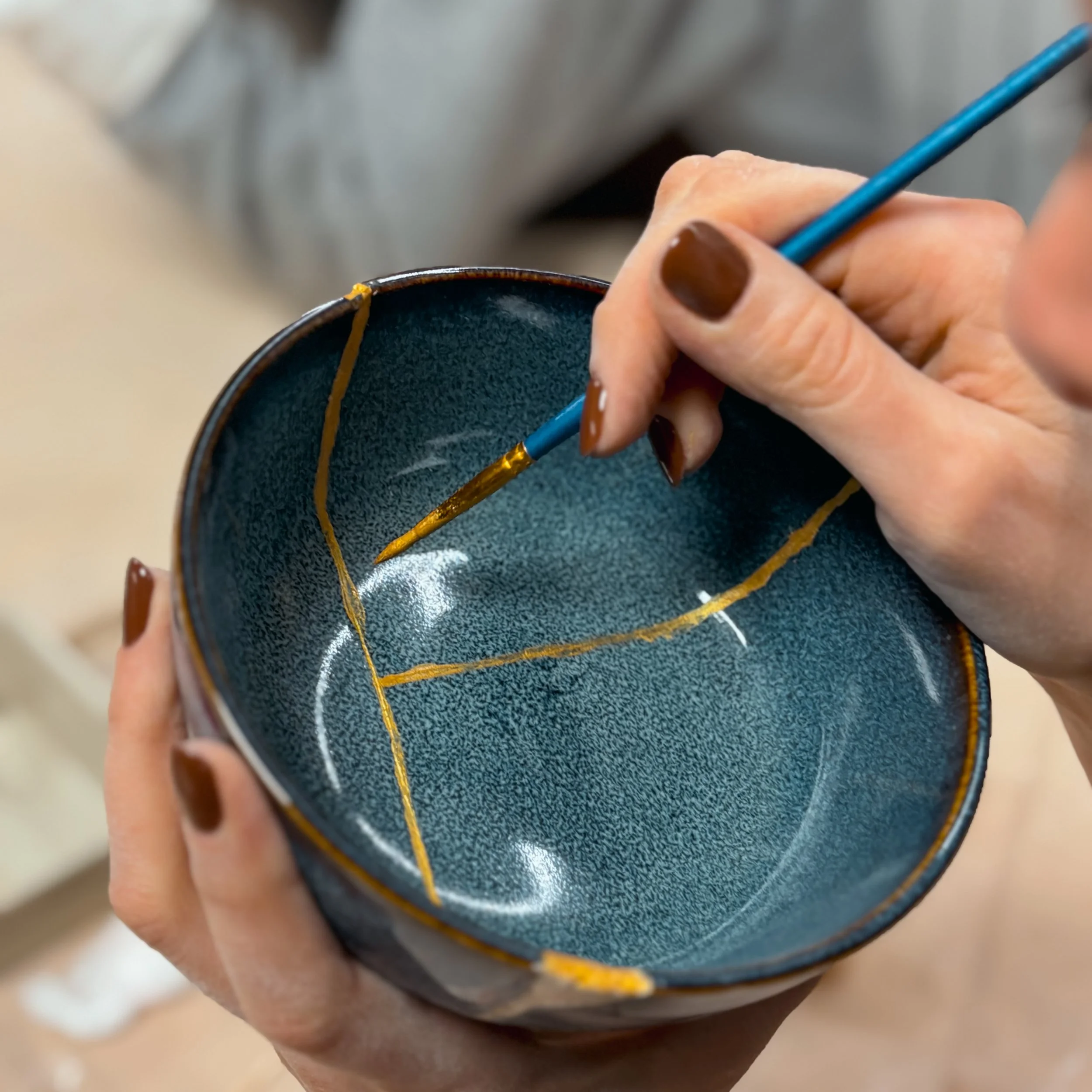 Una persona sta dipingendo un sottobicchiere di ceramica blu con dettagli dorati utilizzando un pennello.