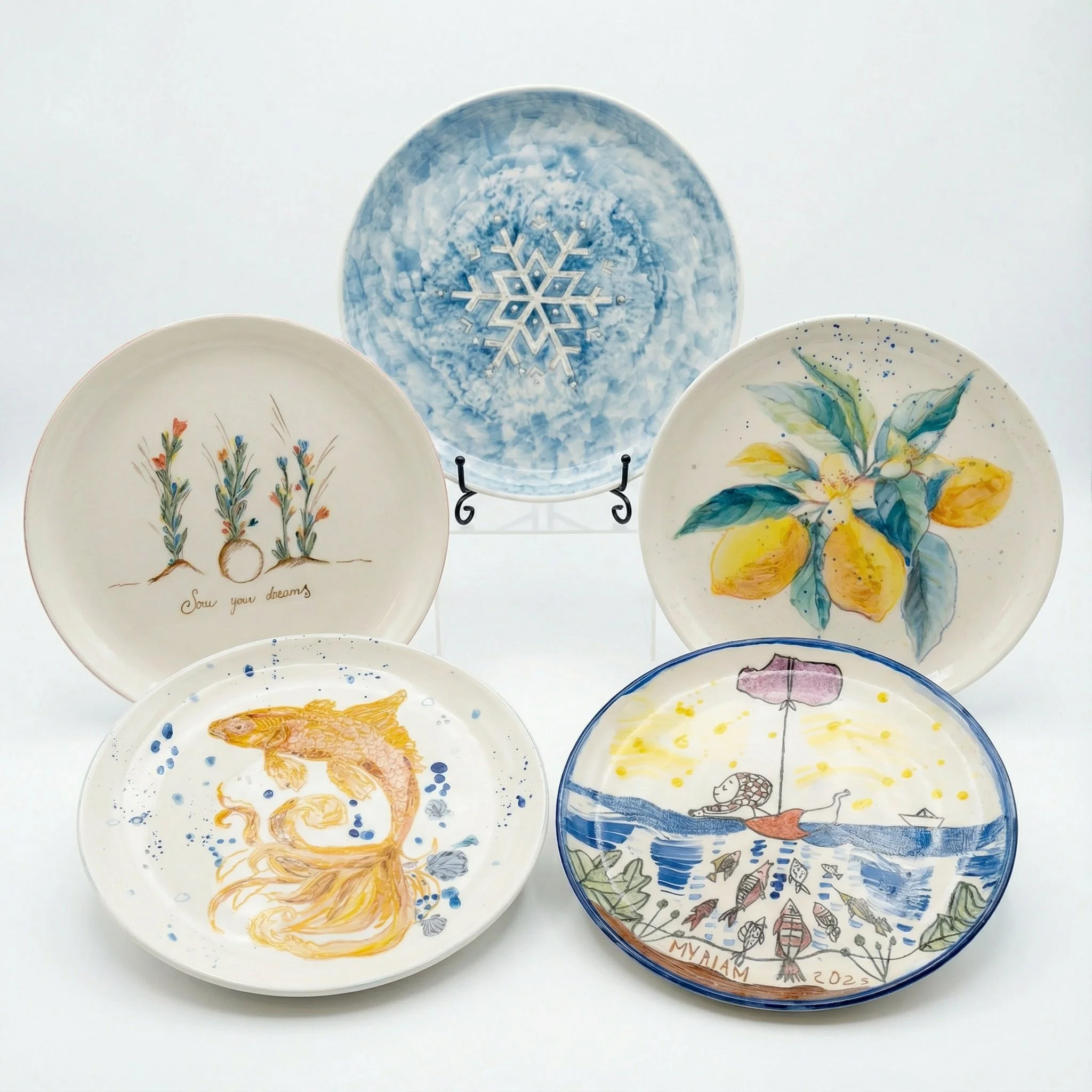 Set di sei piatti decorativi con motivi artistici e colorati, tra cui un fiore di limoni, un pesce, un'onda, un ombrello e una scena marina con pesci e una barca, su uno sfondo bianco.