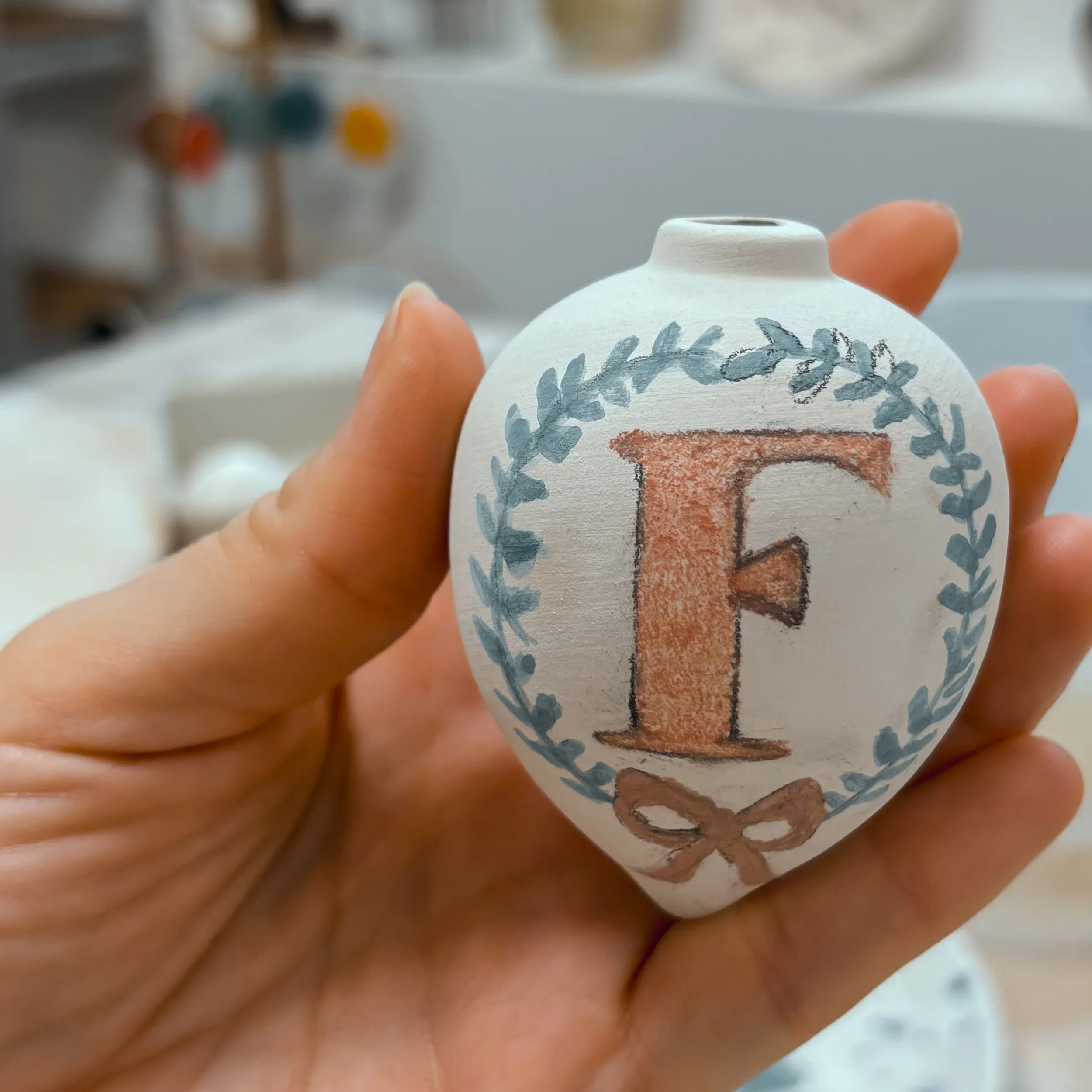 Vaso di ceramica bianco decorato con una lettera 'F' color marrone, contornata da una corona di foglie blu e un fiocco marrone alla base.