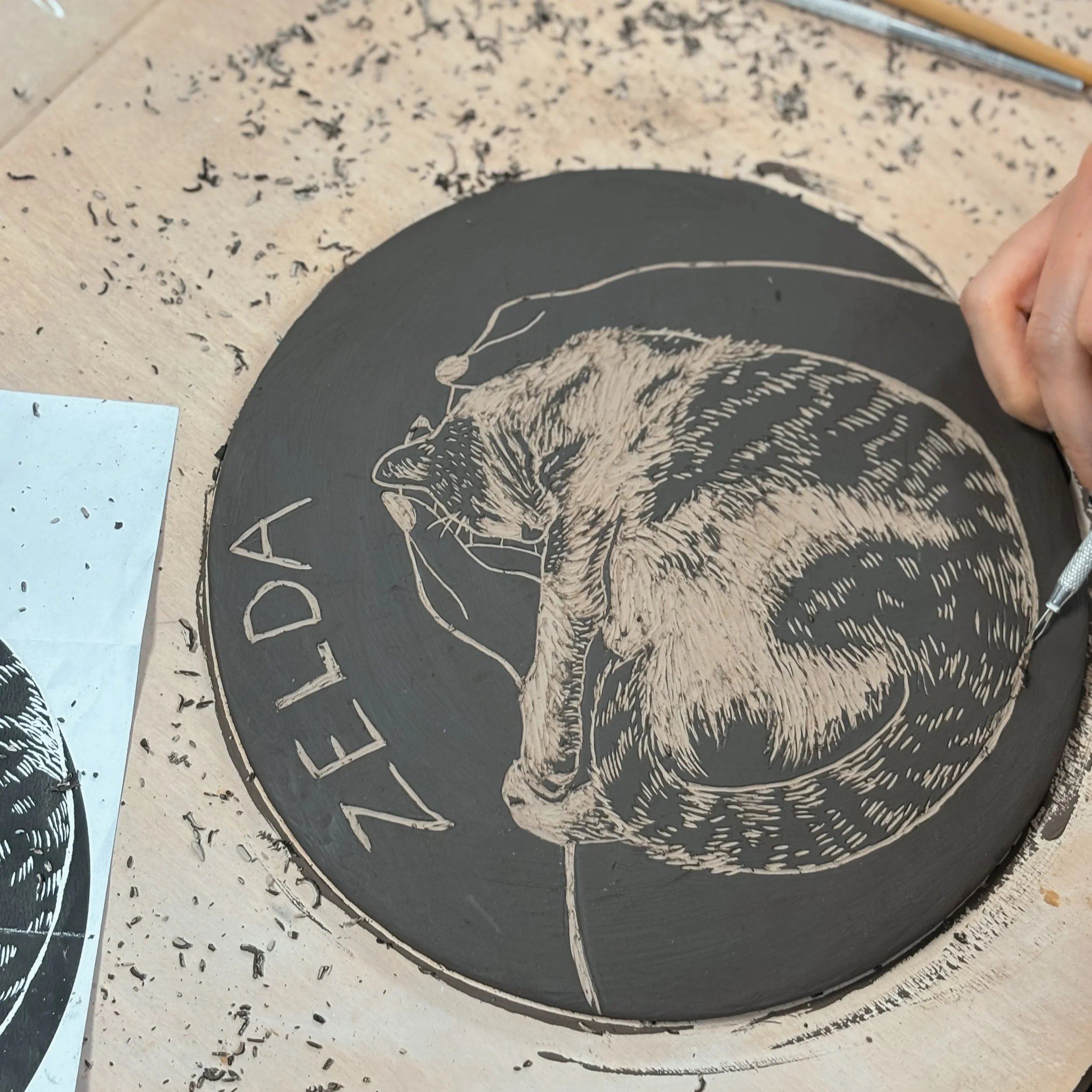 Una persona sta scolpendo un'incisione di un gatto su un disco di pietra nero, con la parola 'ZELEDA' scritta a fianco, utilizzando strumenti di incisione.