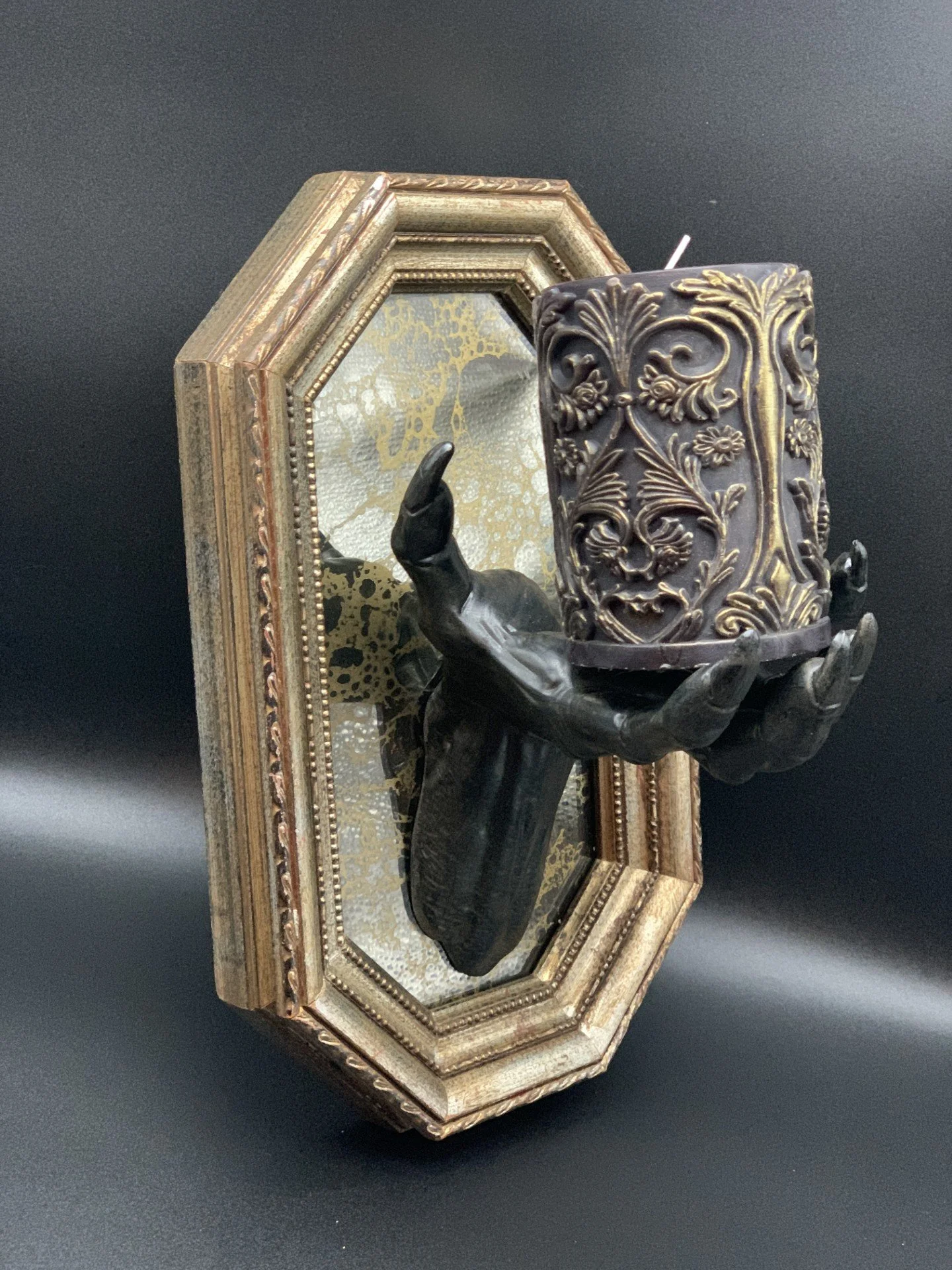 A black hand sculpture holding an ornate decorated candle holder with a large candle, mounted on a decorative gold frame mirror.
