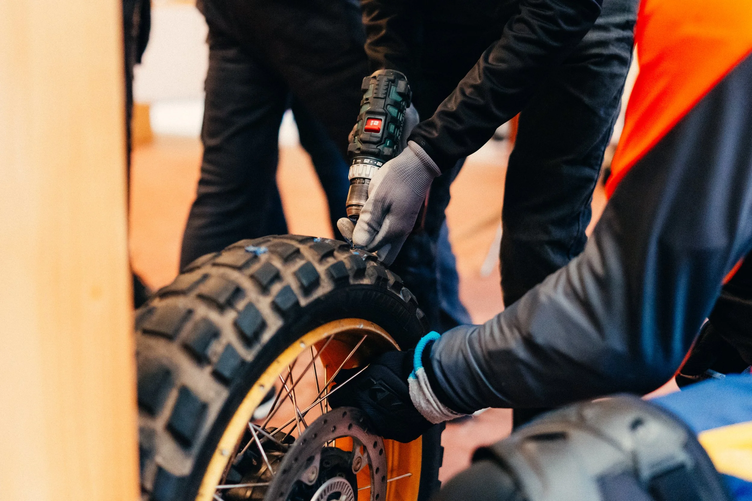 Two individuals working on a motorcycle tire, one using a cordless drill, both wearing gloves and black clothing, with a person in an orange and black jacket helping.