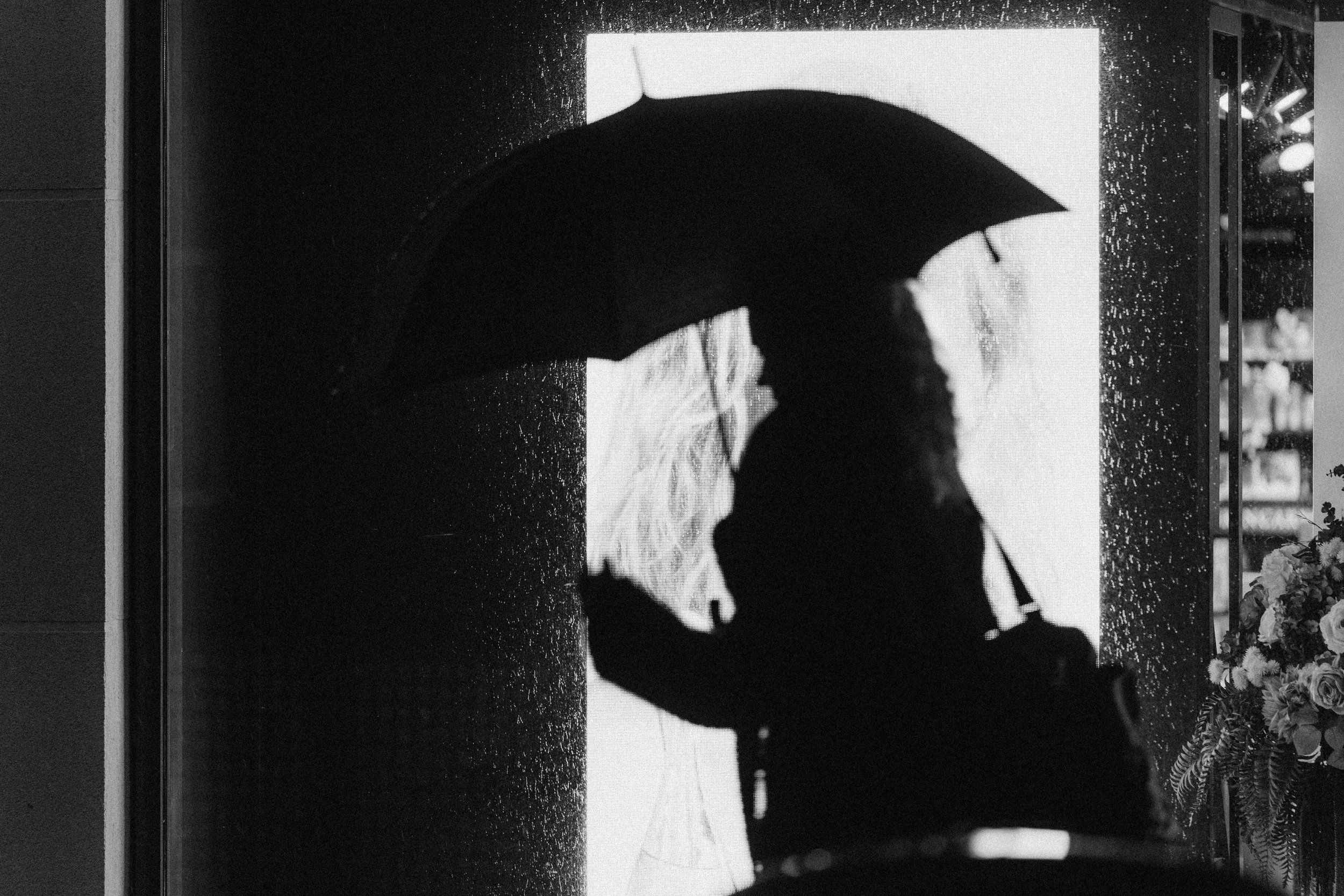 Silhouette of a person holding an umbrella on a rainy day, standing near a lit display case with flowers to the right.