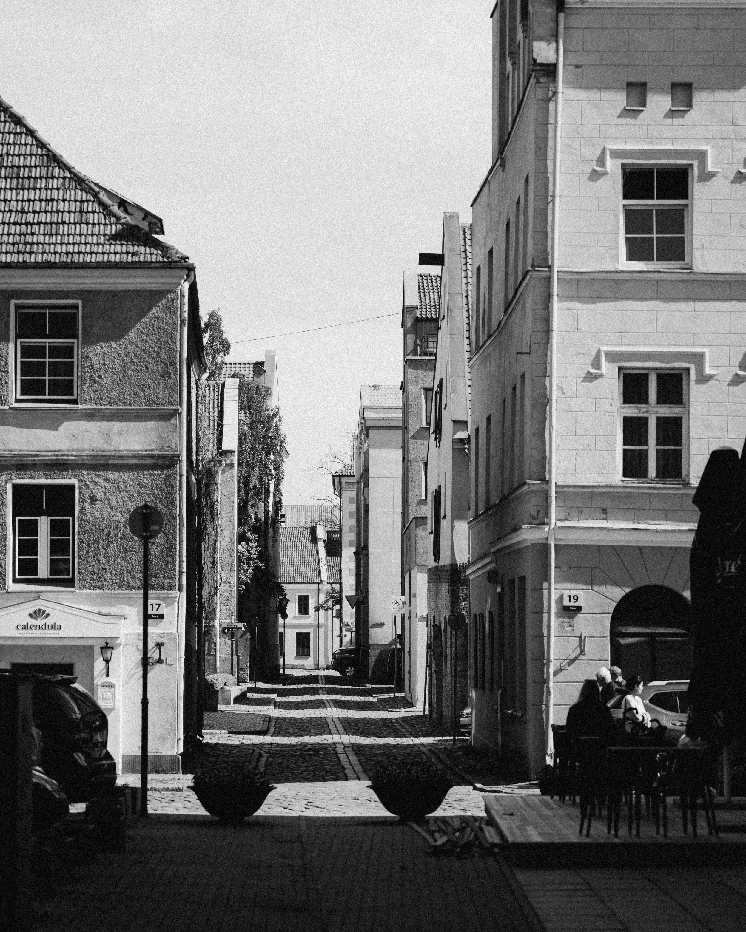 Black and white photo of a narrow city street with old buildings, parked cars, and people sitting at outdoor tables.