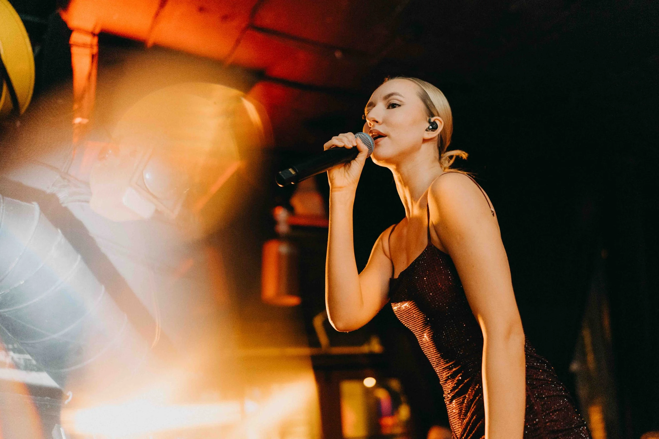 A woman singing into a microphone on stage, wearing a sparkly black dress with a tattoo on her left shoulder, and a wireless earpiece, with warm stage lighting and dark background.