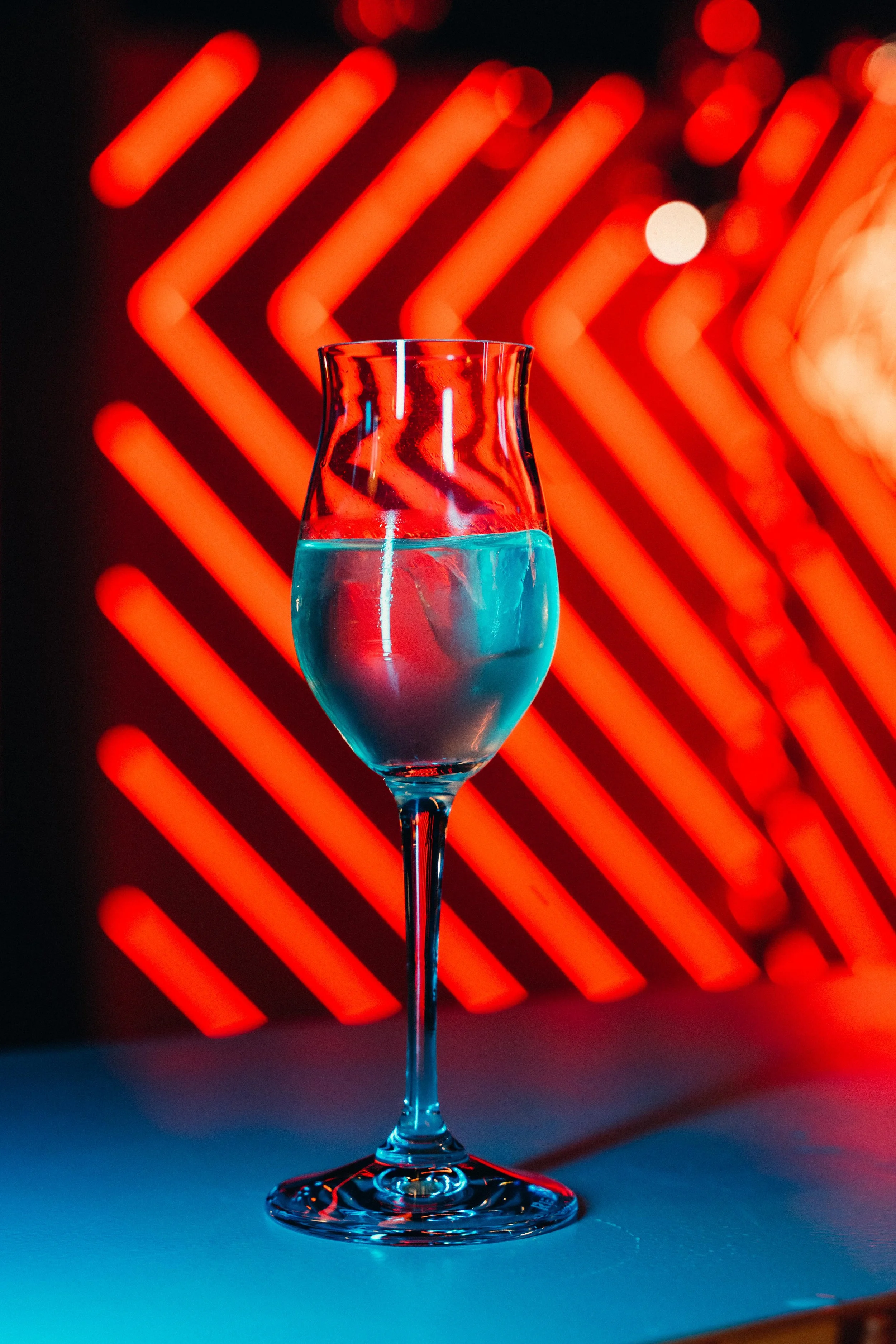 A tall glass with a long stem and curved bowl, filled with a blue liquid, set against a background of red neon lights forming chevron patterns.