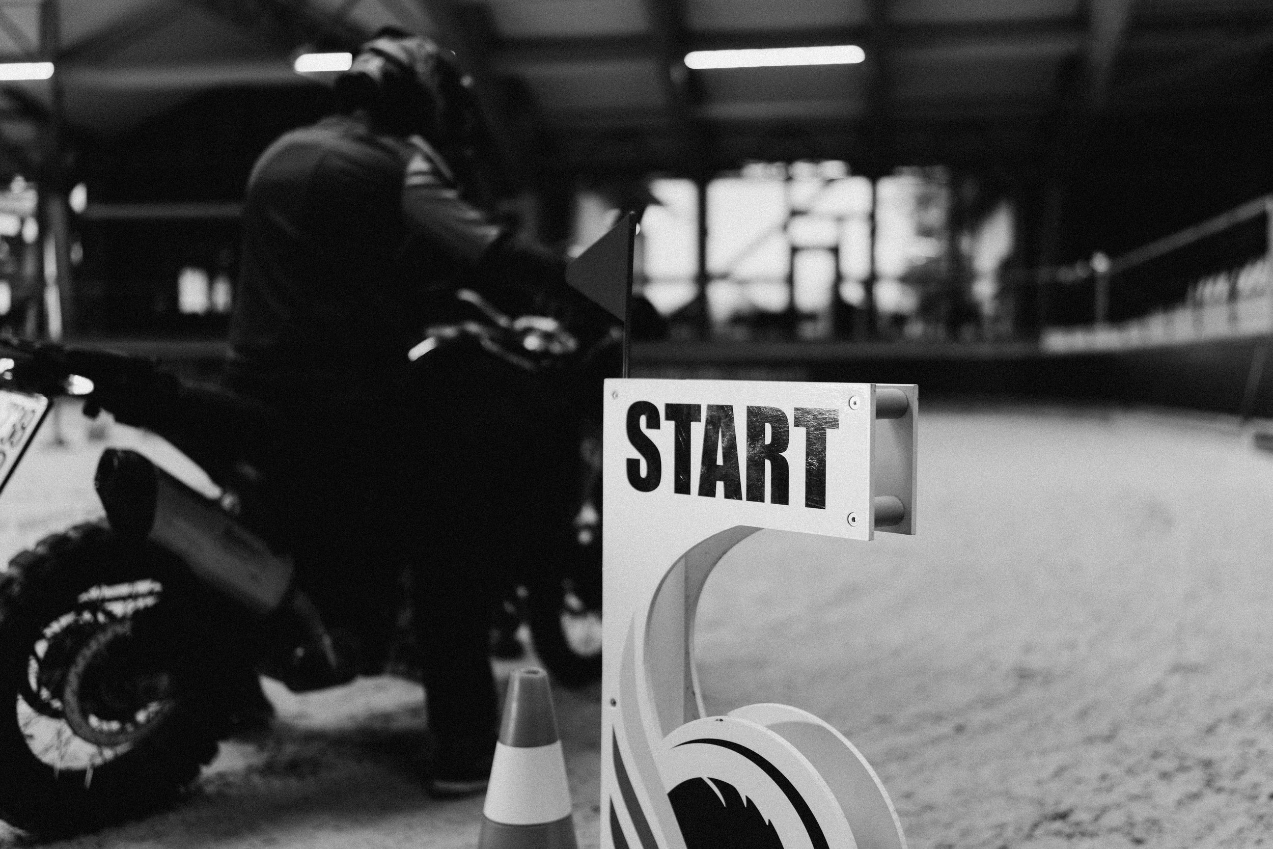 A motorcyclist preparing to start a race at a gated start line with a 'START' sign and a traffic cone in the foreground, inside a large indoor arena.