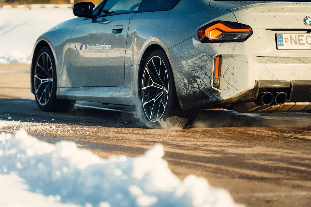 A silver BMW car driving on a snow-covered road, kicking up snow behind the rear tires, with visible dirt and mud on the back bumper. The car has a license plate from Lithuania.