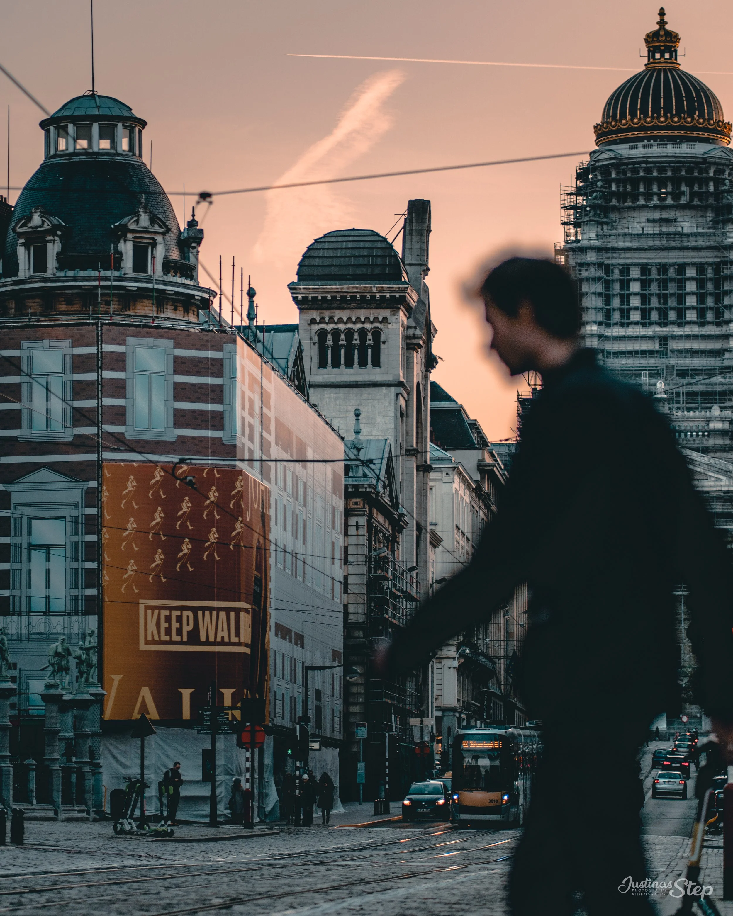 A person walking in an urban street during sunset, with historic buildings and construction scaffolding, and a large sign that says 'Keep Walking'.