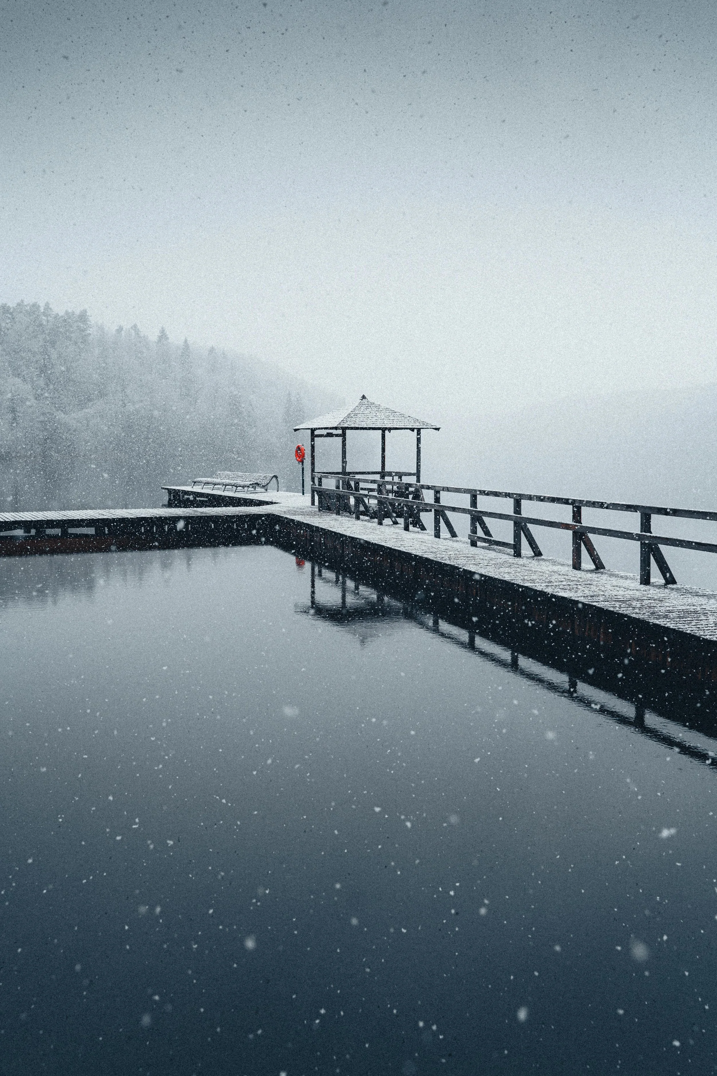 Snow-covered wooden pier with gazebo and snow falling over calm water, foggy landscape in the background.