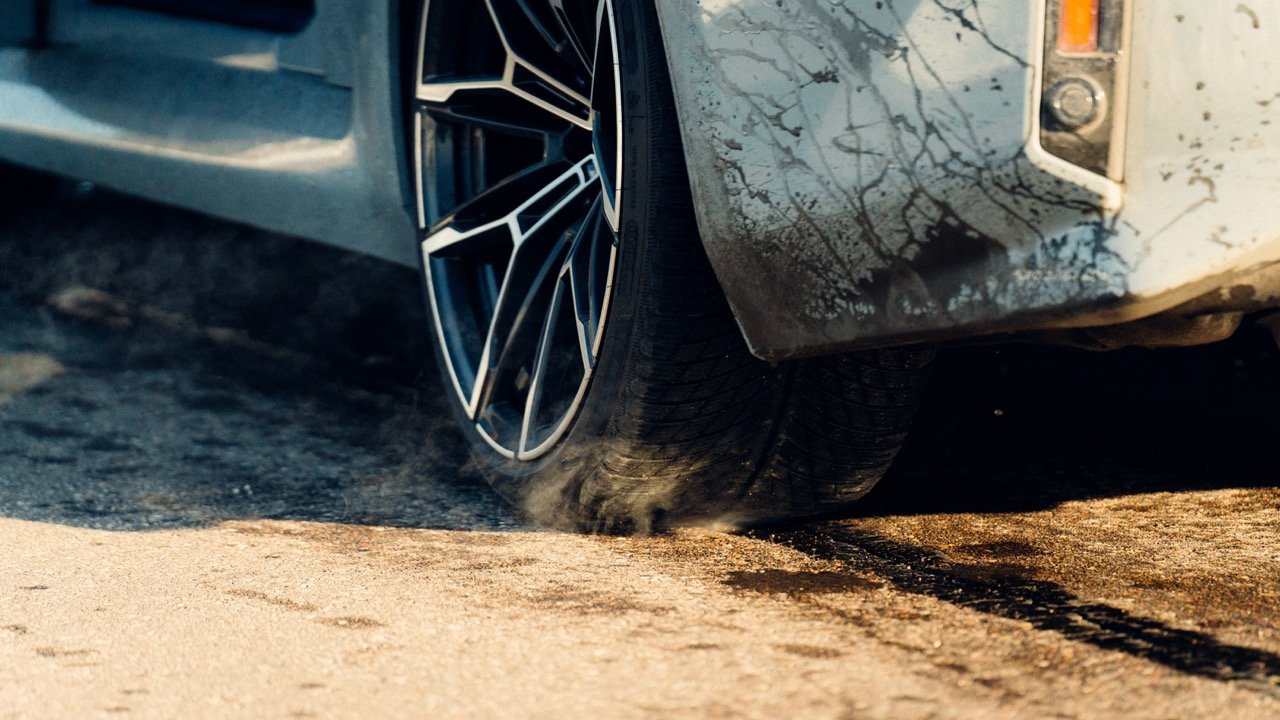 Close-up of a car tire rolling over a dirt and asphalt surface, with dust and dirt visible around the tire.