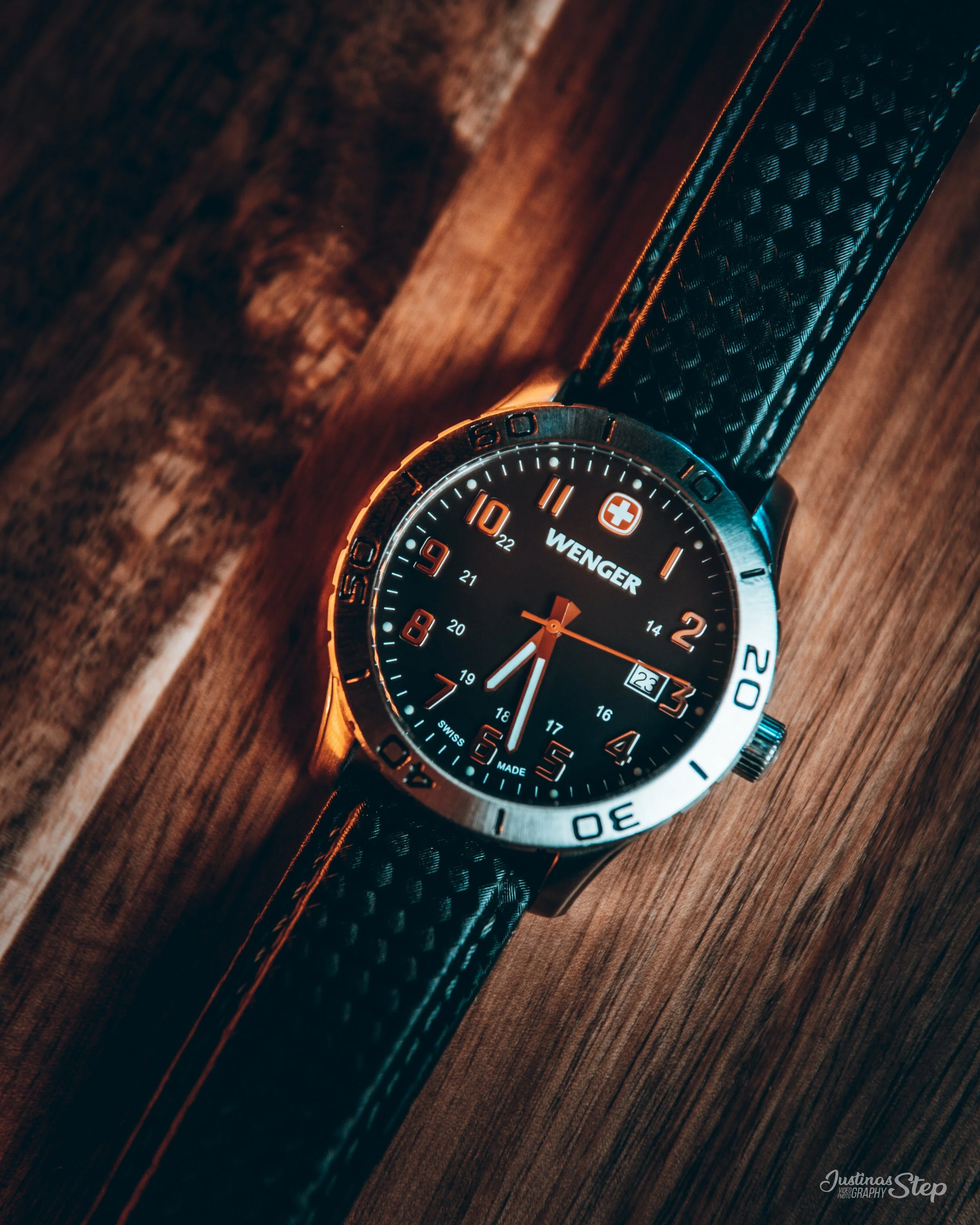 A wristwatch with a black face, silver bezel, and black textured strap resting on a wooden surface.
