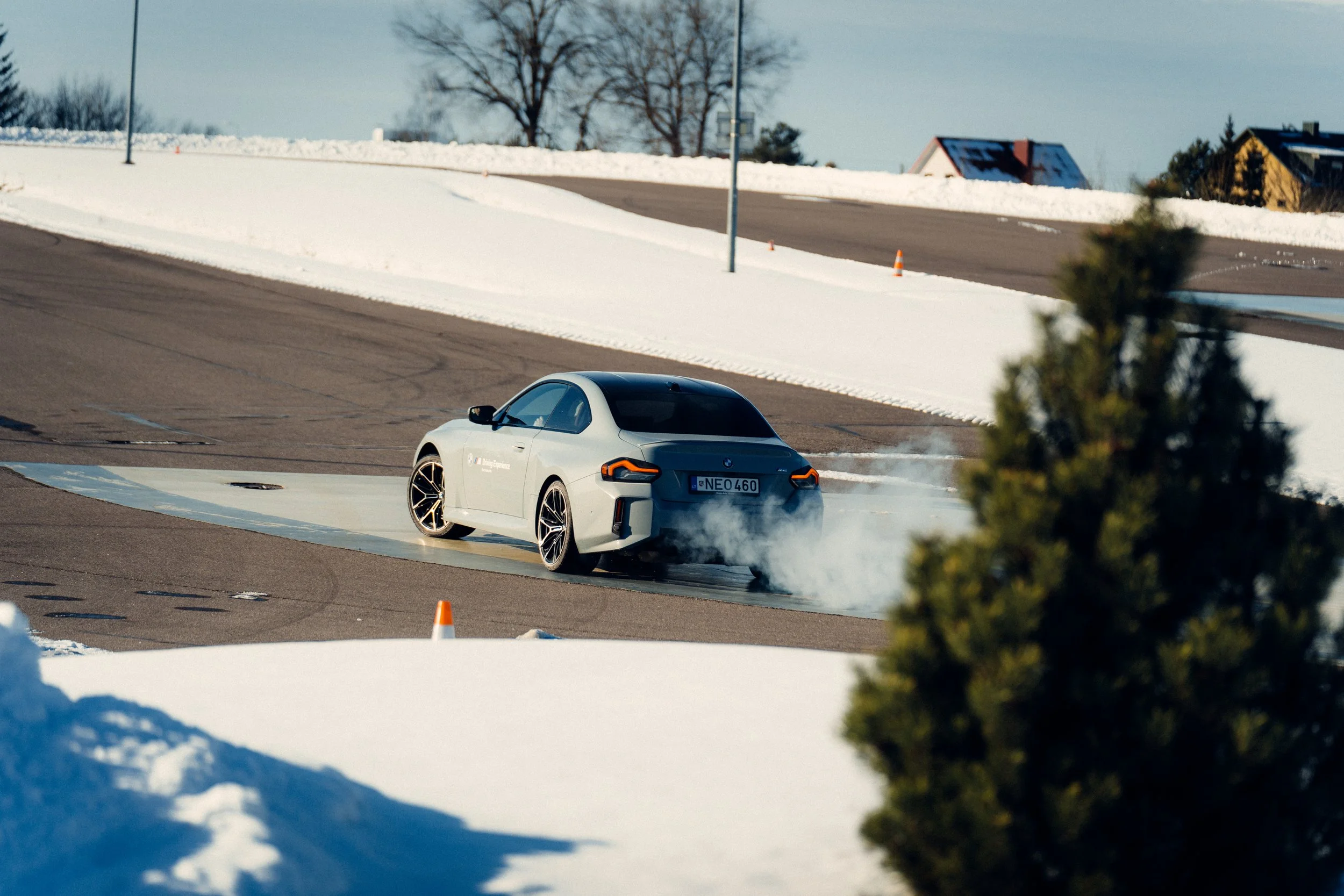 A white BMW car performing a burnout on a wet, snow-covered road in a winter setting with snow on the ground and trees in the background.
