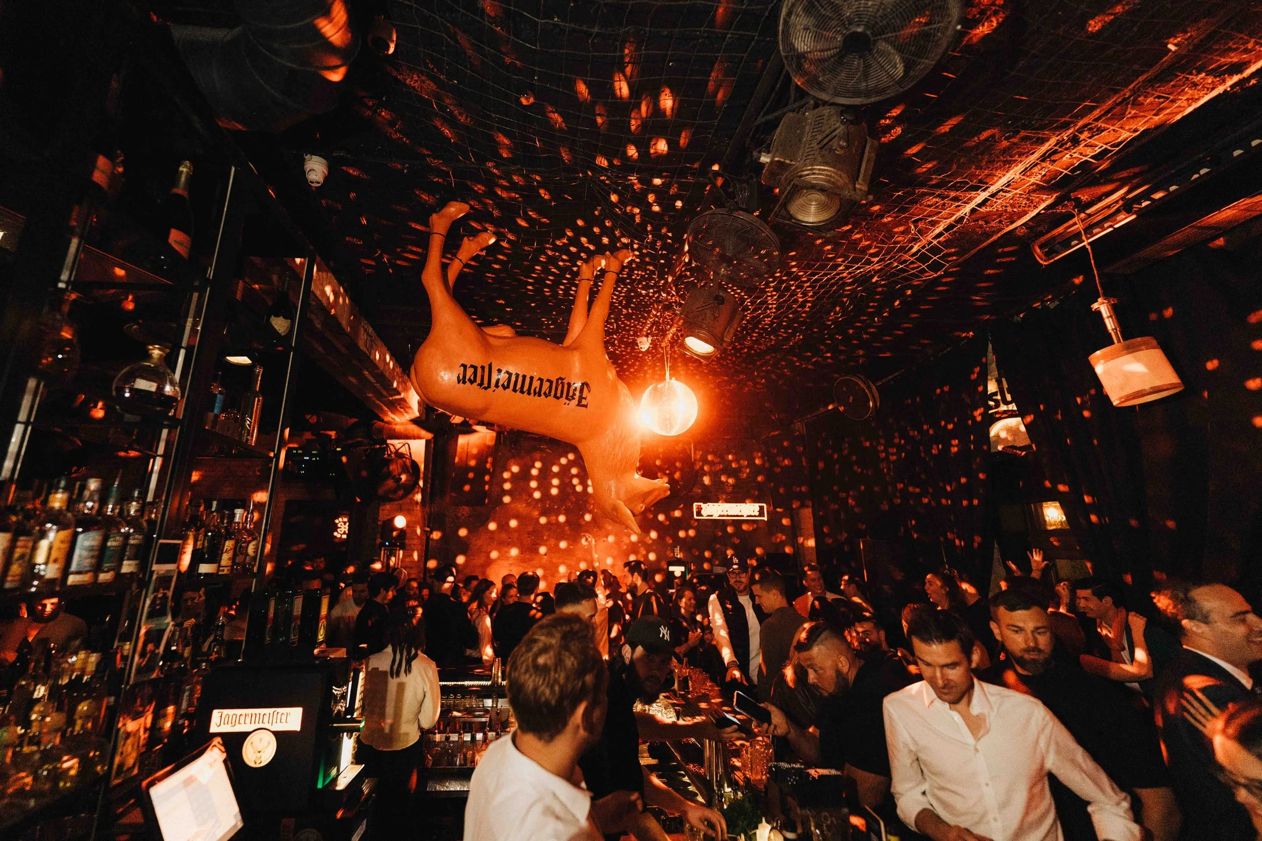 A crowded nightclub scene with people dancing and socializing. A large inflated pig with the word 'Jägermeister' written on it hangs from the ceiling, illuminated by red and orange lighting, creating a festive atmosphere.