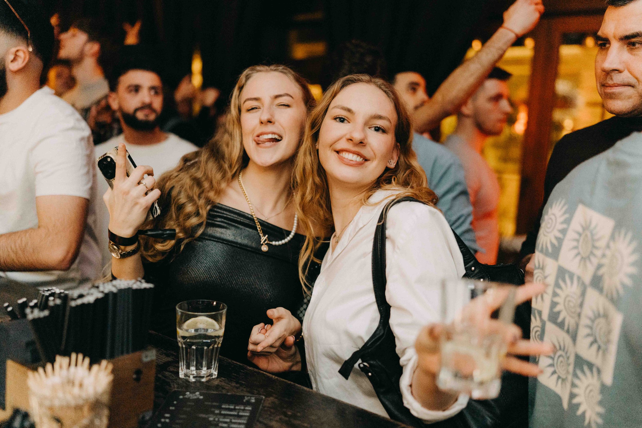 Two young women with curly red hair smiling and having fun at a crowded bar or party, holding drinks, with other people in the background.