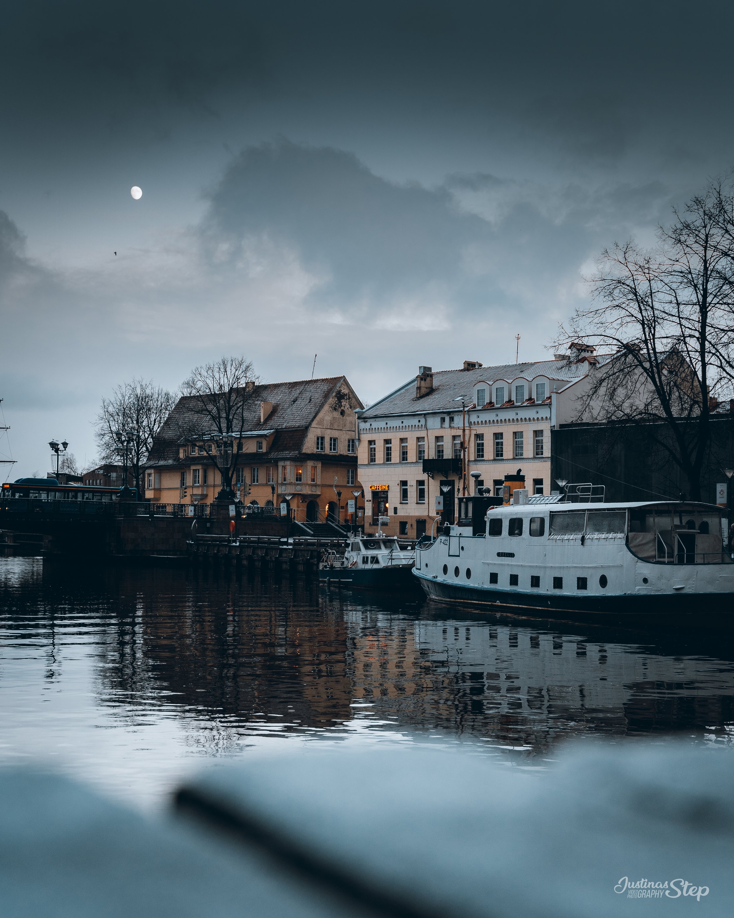 A harbor scene at dusk with boats floating on calm water, overcast sky with dark clouds, a visible moon, and historic buildings along the waterfront.