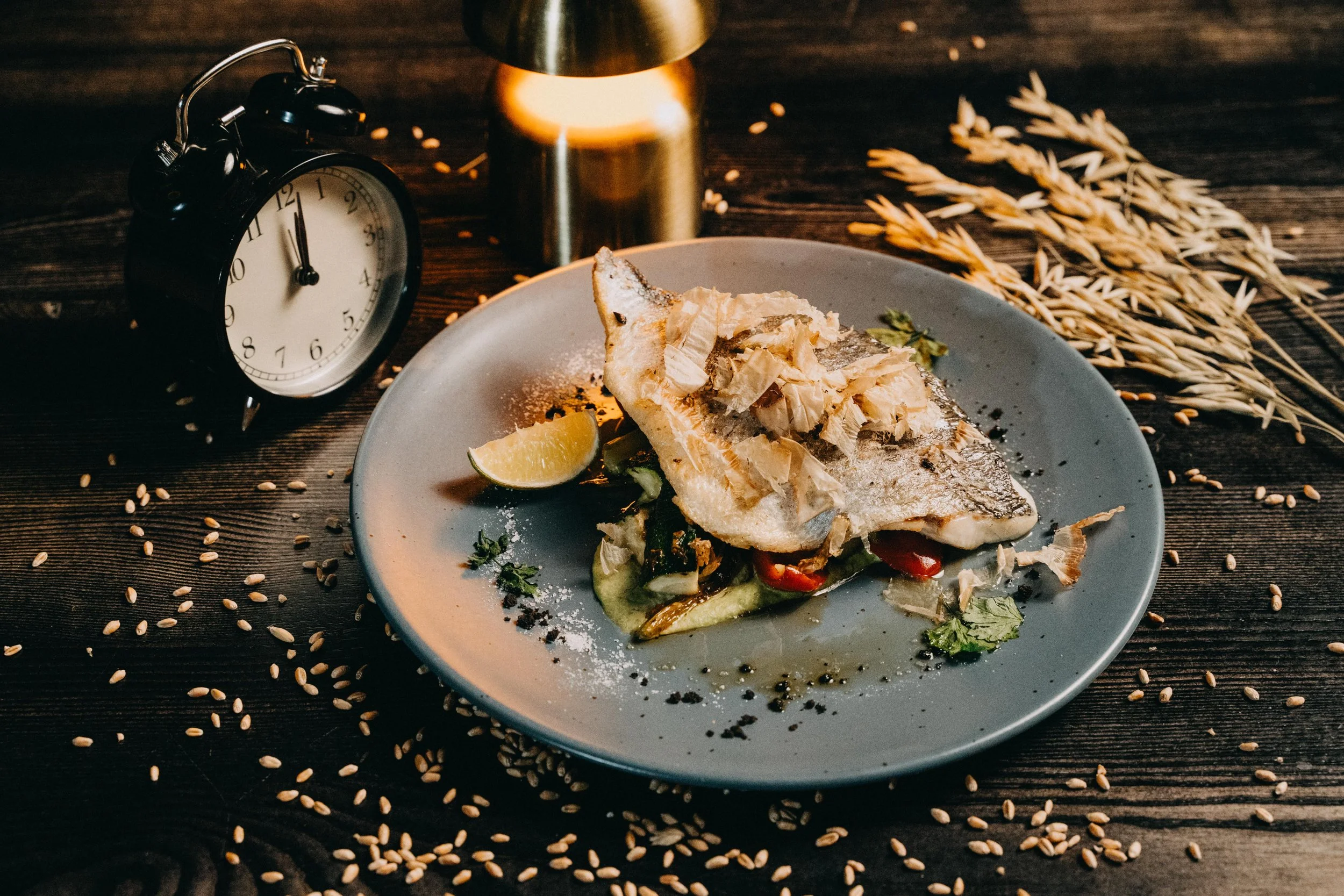 A plate of cooked fish garnished with shredded toppings, lemon wedge, and vegetables on a rustic wooden table, with a black alarm clock, a candle, and sprigs of dried wheat around it.