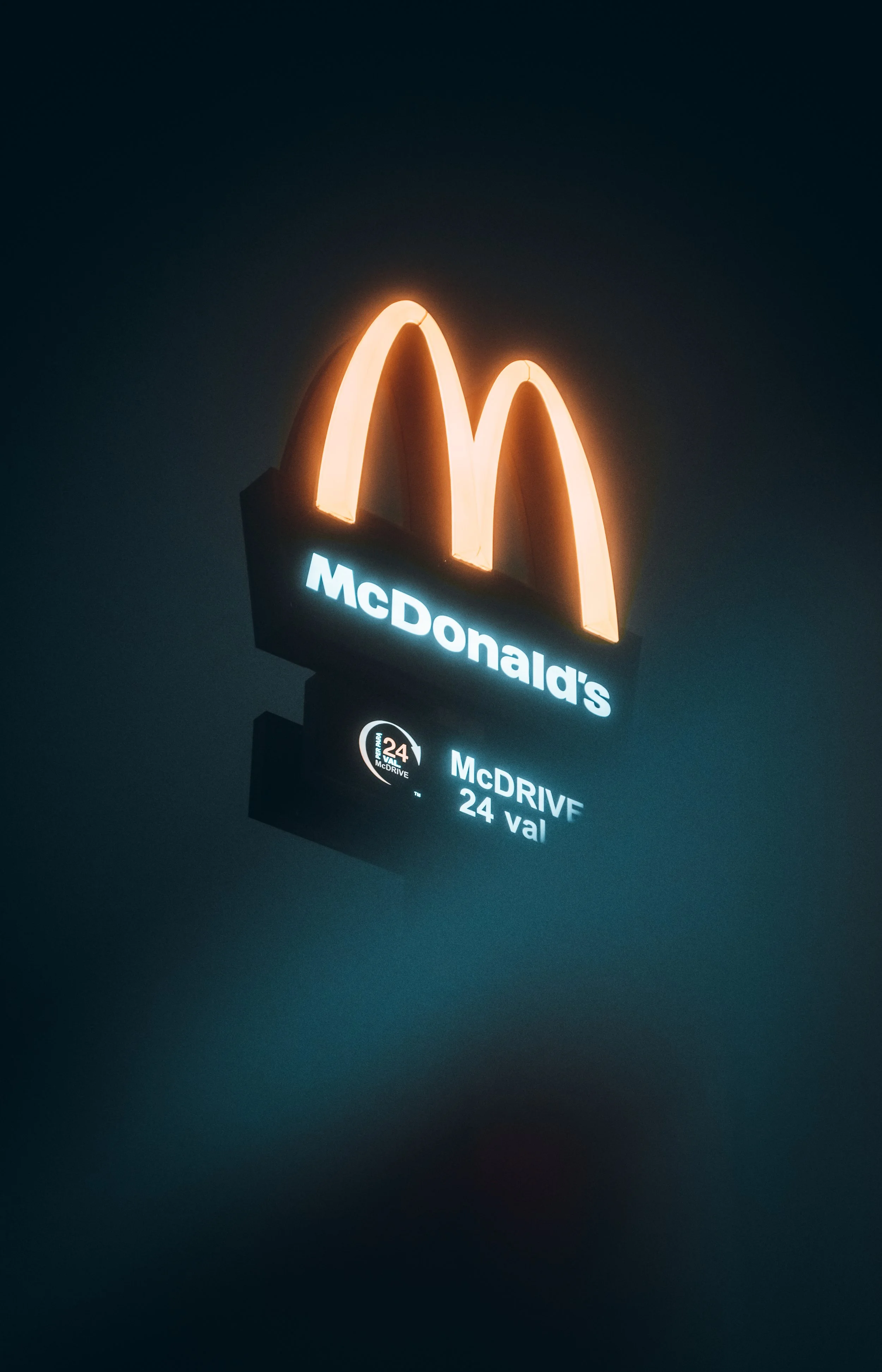 A neon sign of the McDonald's logo with illuminated golden arches and the text 'McDonald's' underneath, along with a smaller digital display below showing 'McDRIVE 24 val' against a dark background.