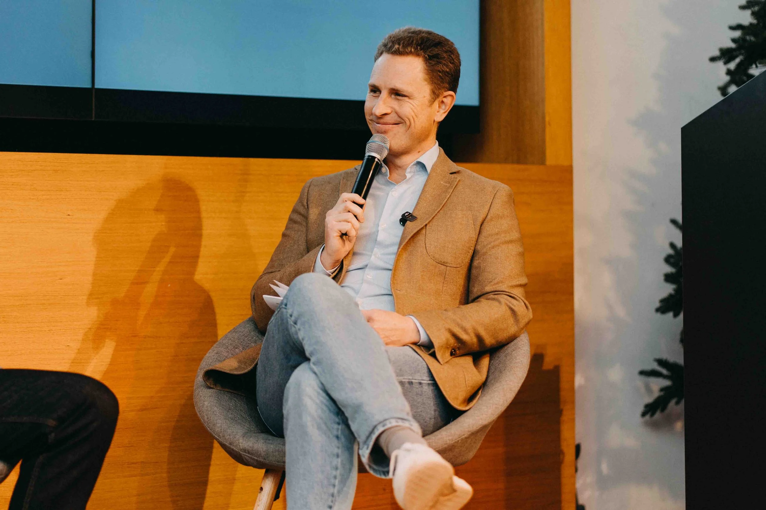 A man in a brown blazer and light jeans sitting in a gray chair, holding a microphone and smiling during a discussion or presentation.