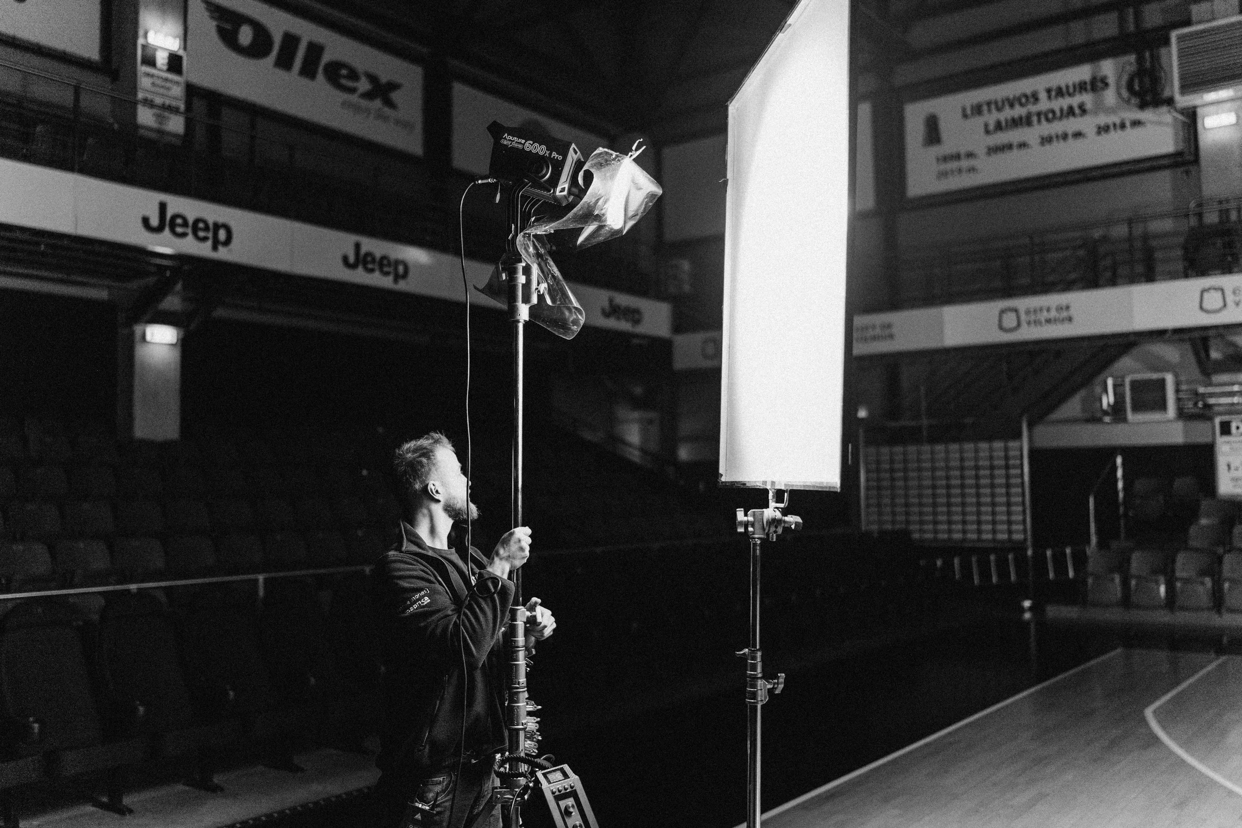 A person working as a photographer in an indoor sports arena, setting up a lighting or camera setup near an empty basketball court, with advertising banners on the walls.