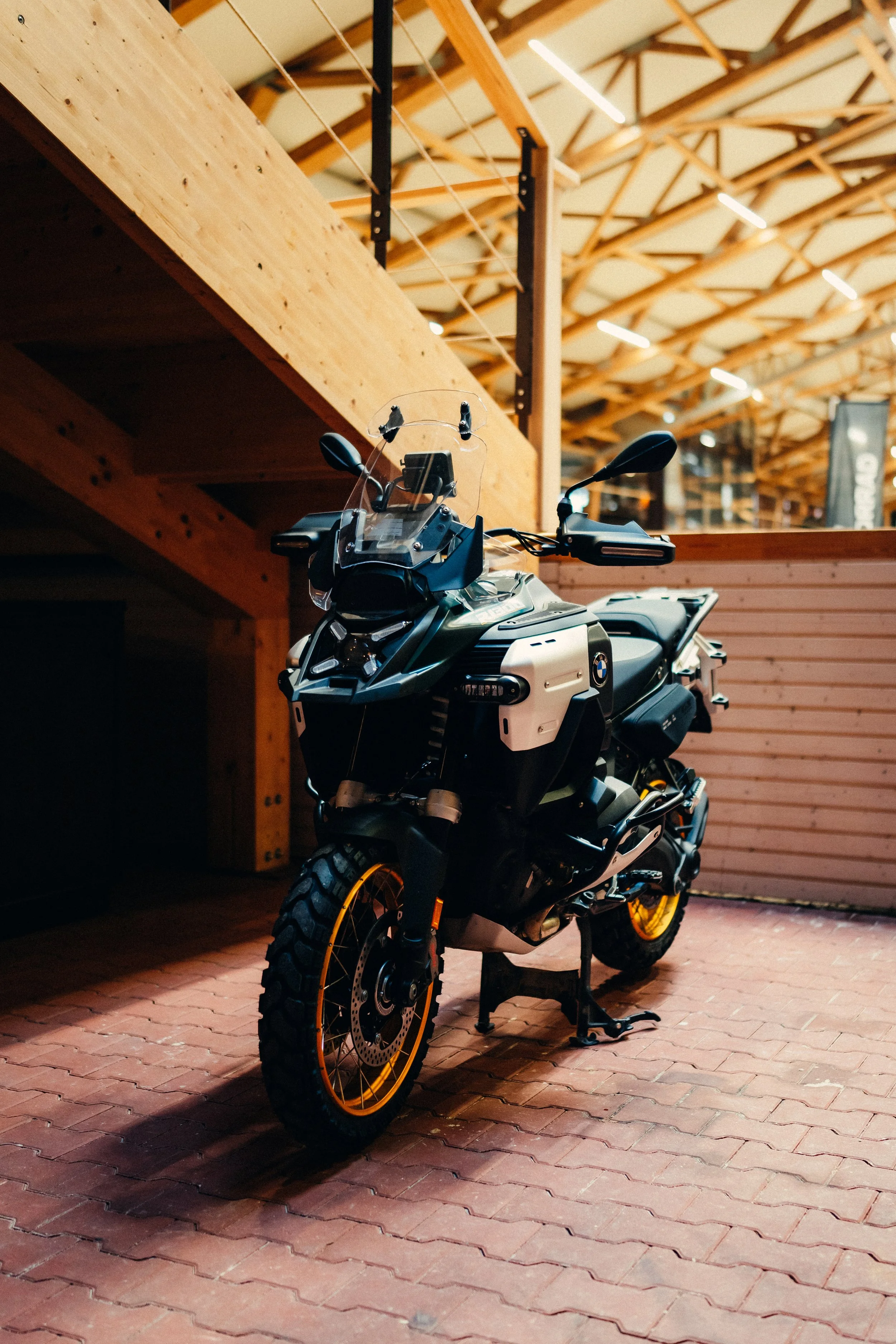 Black adventure motorcycle with yellow rims parked indoors, with wooden walls and ceiling structure.