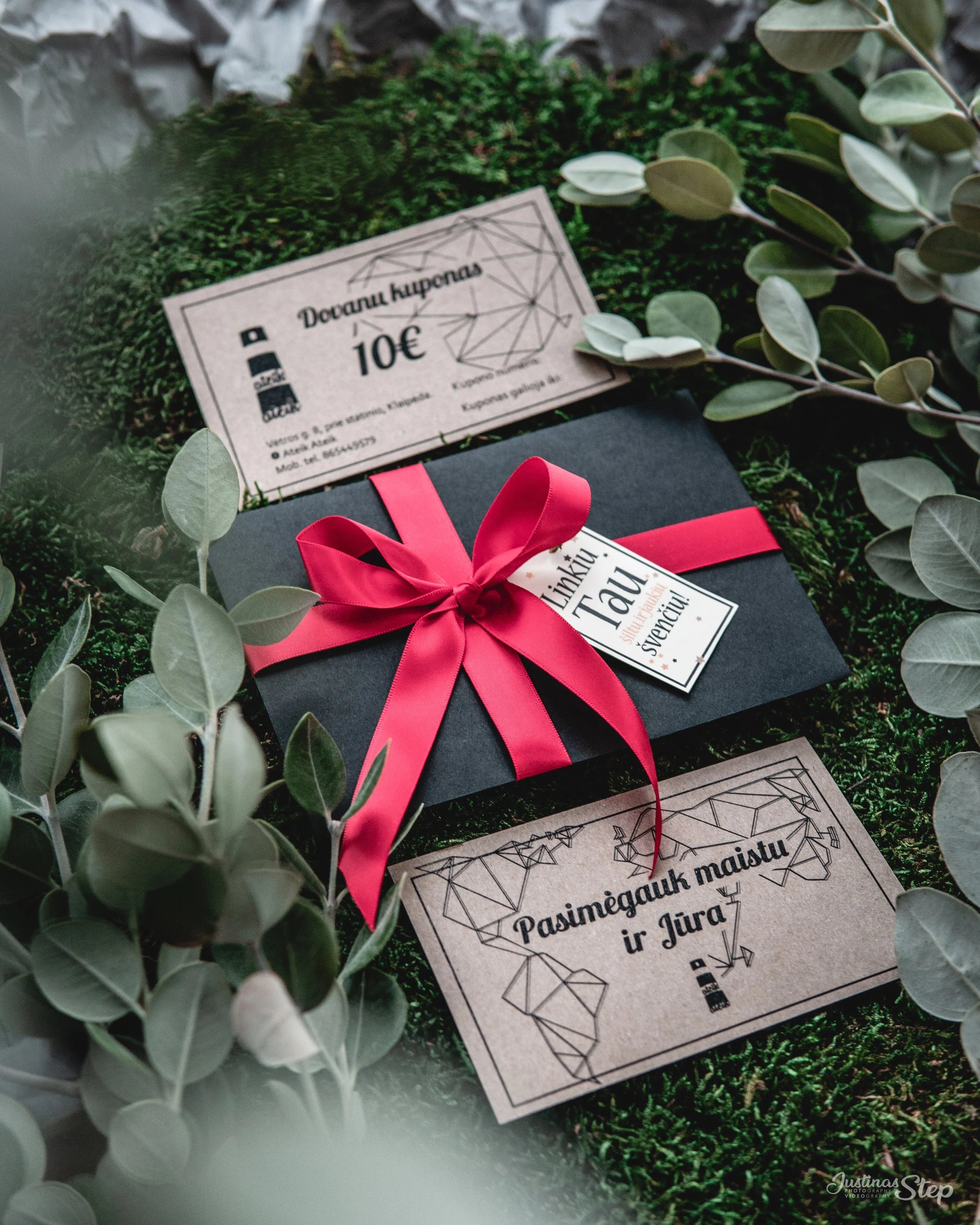 A black gift box with a pink ribbon bow, surrounded by greyish green leaves, positioned on a mossy green surface. There are Lithuanian coupons and a greeting card along with a price tag nearby.