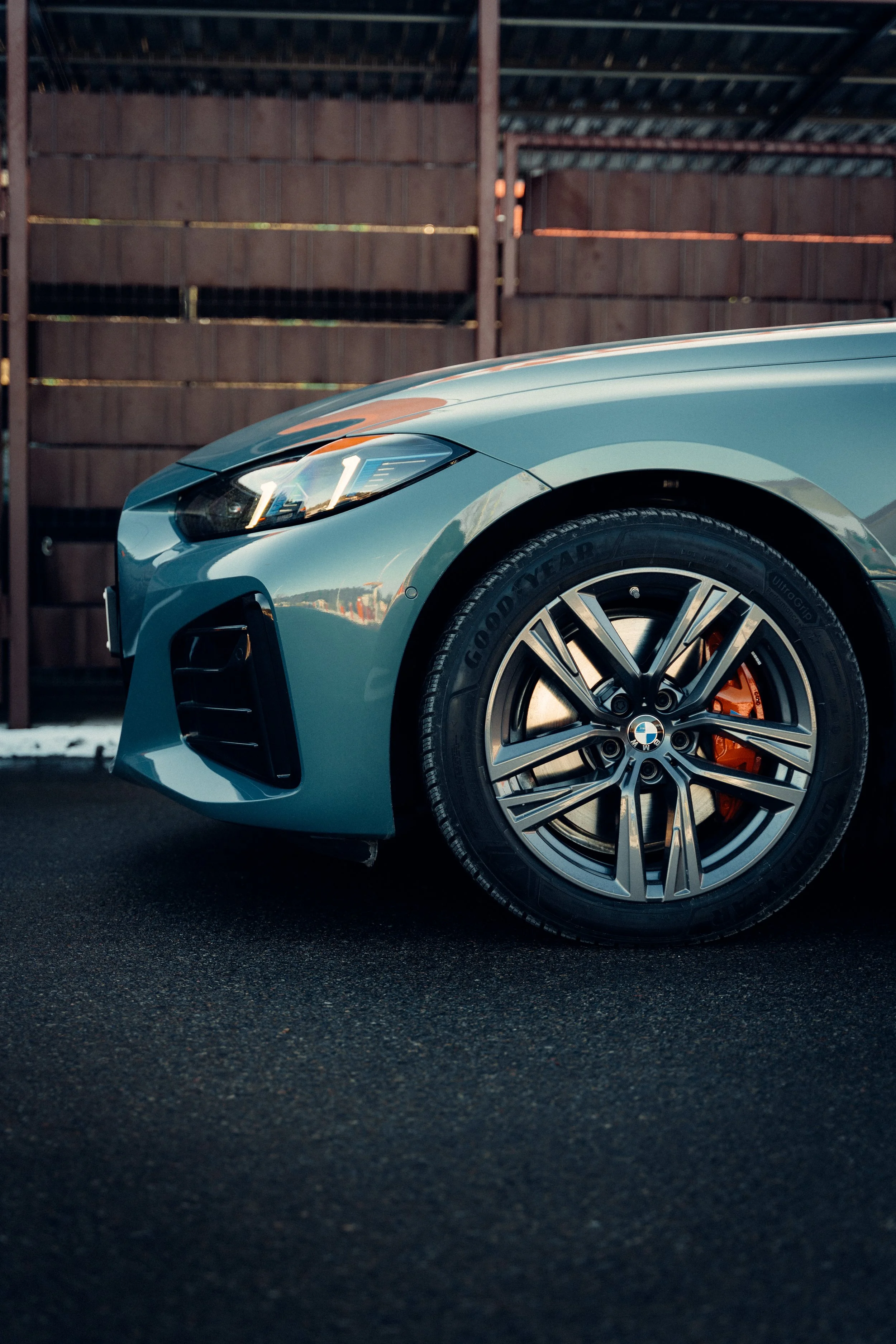 Close-up of the front left side of a blue BMW car parked on a paved surface, showing the wheel with a BMW logo and orange brake caliper, against a wooden fence background.