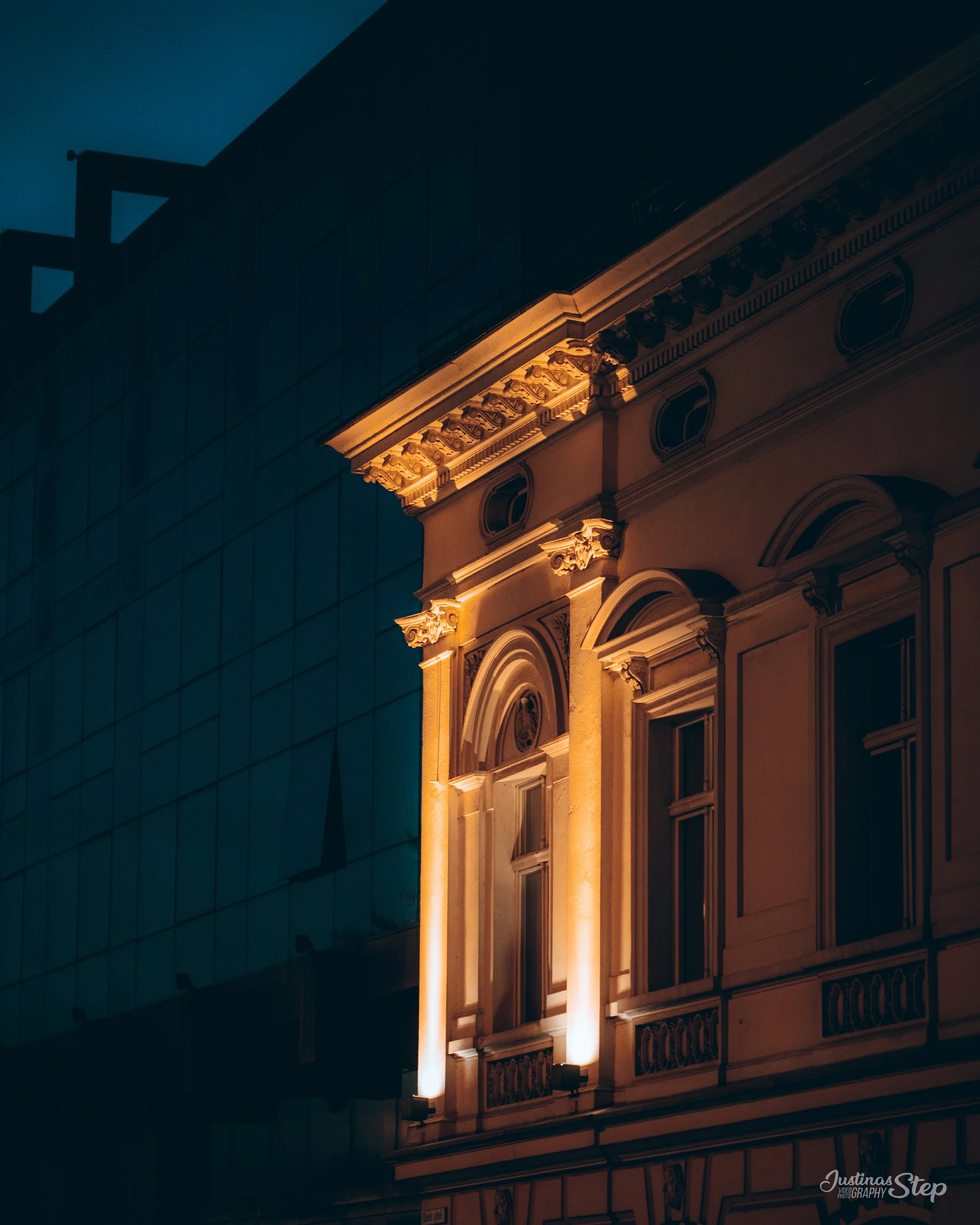 Night view of an ornate building with classical architectural details illuminated by warm exterior lighting, adjacent to a modern glass building.