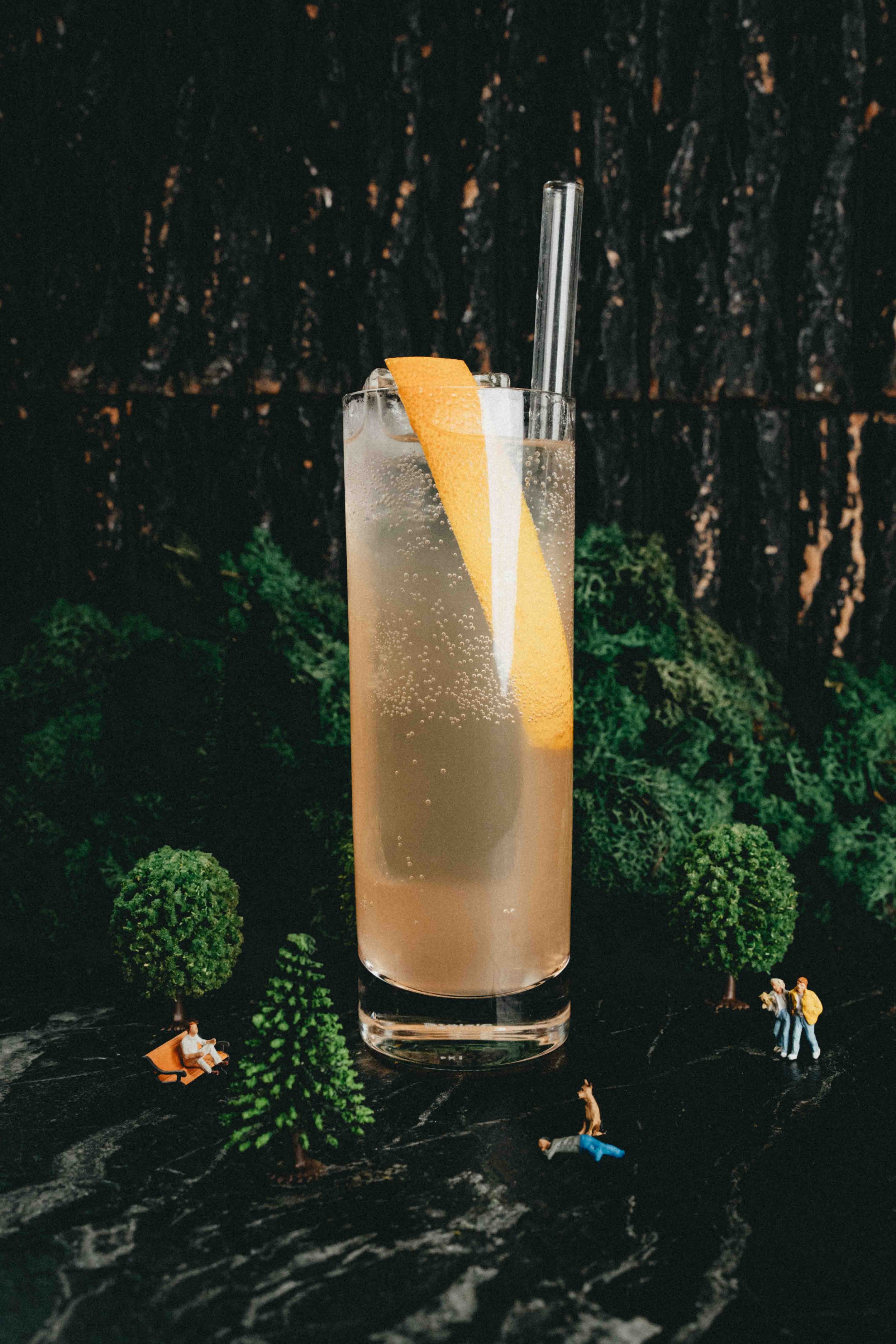 A tall glass of a sparkling drink with an orange wedge and a straw on a dark surface with miniature figurines and trees creating a miniature park scene.