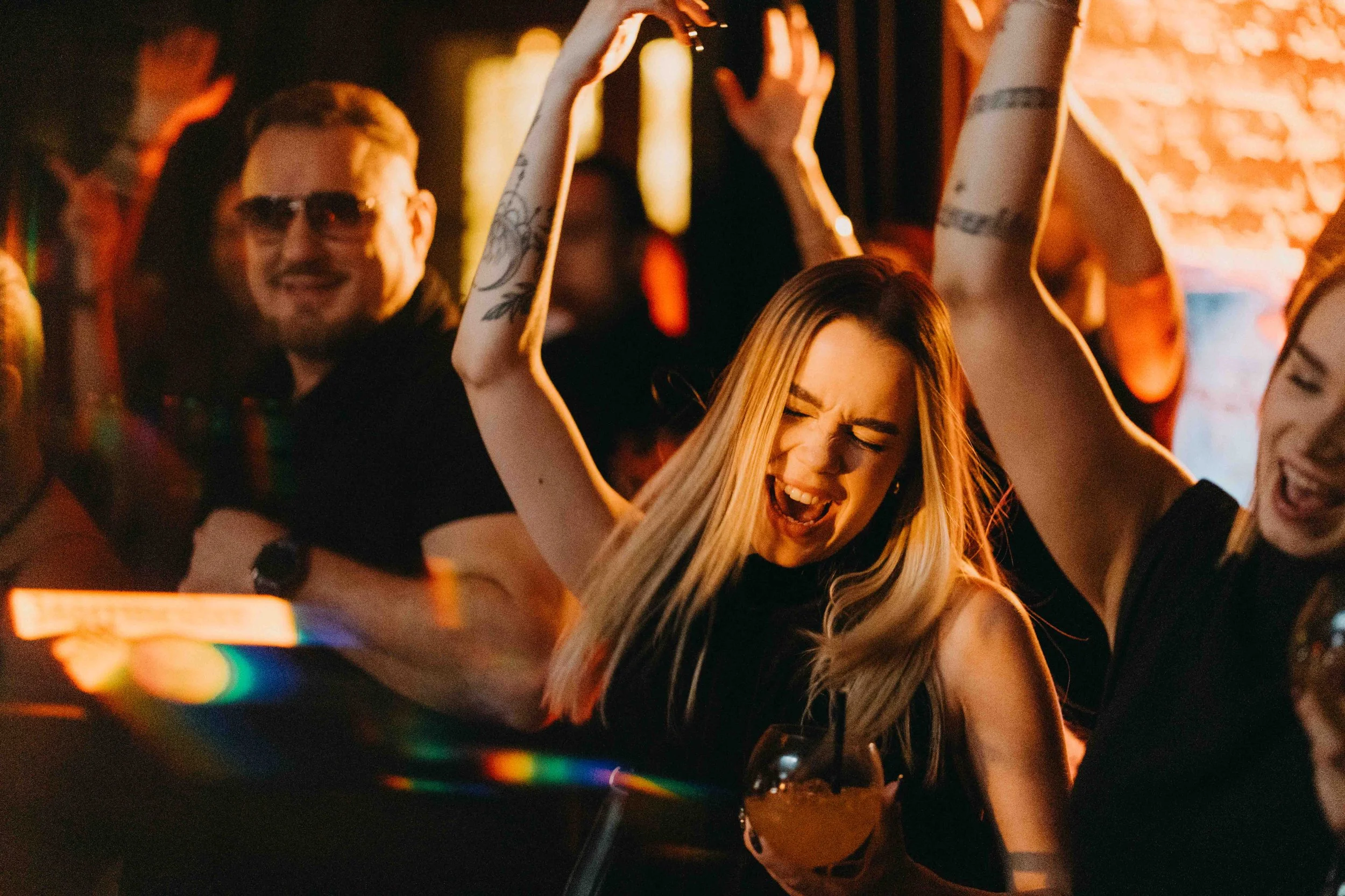 People dancing and singing with smiles at a party or nightclub, with warm ambient lighting and a woman in the center holding a drink.