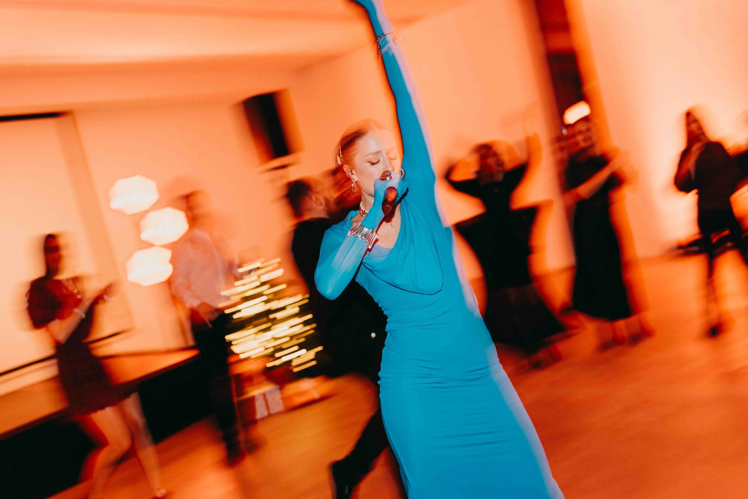 A woman in a blue dress singing into a microphone at a party or event, with people dancing and socializing in the background.