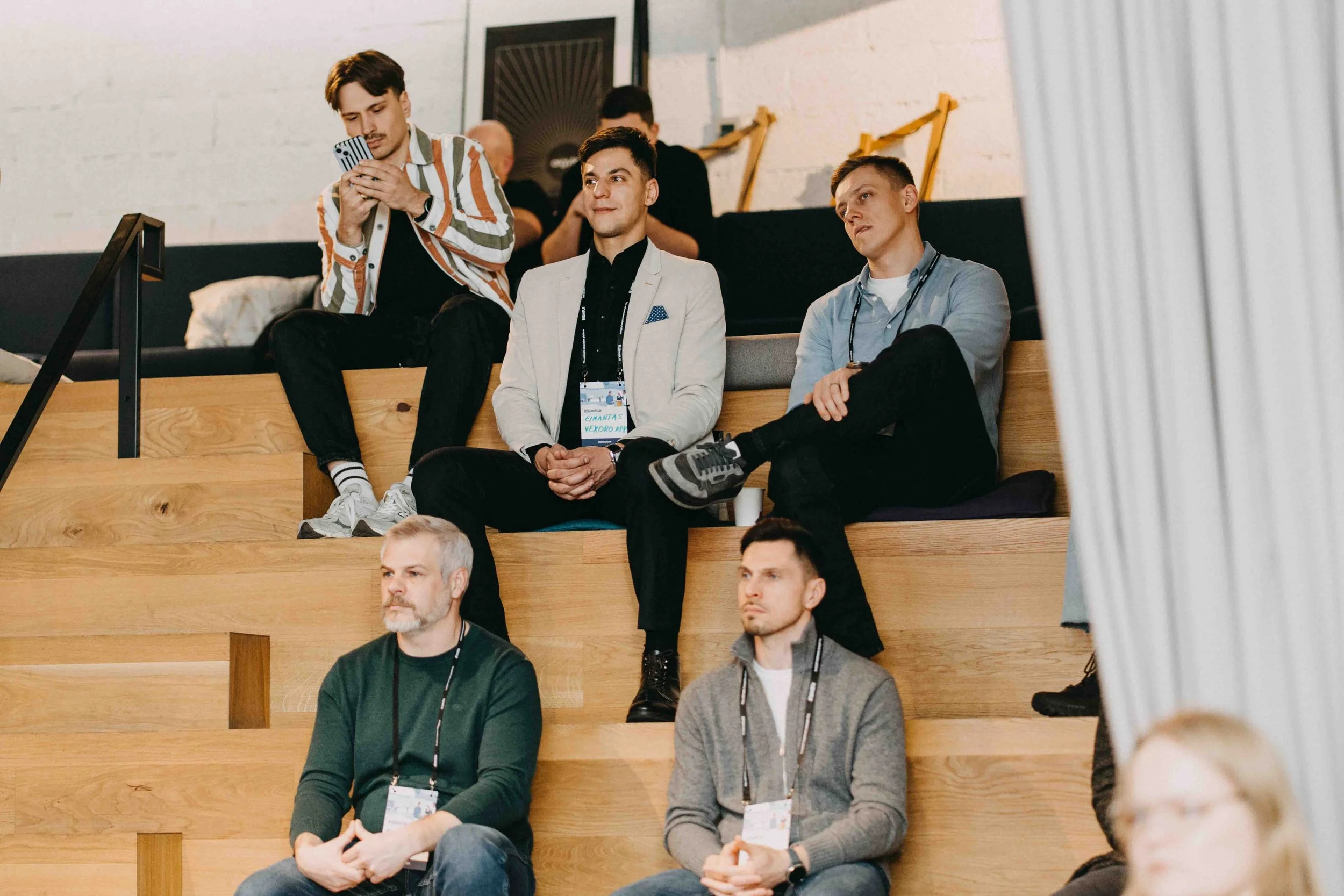 Group of six men sitting on tiered wooden seating in a conference room, some wearing conference badge lanyards, attending a presentation or event.