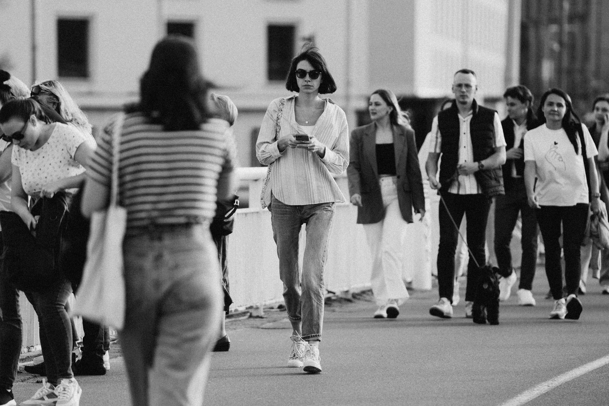 A young woman with short hair and sunglasses walking while looking at her phone among a group of people on a city street.
