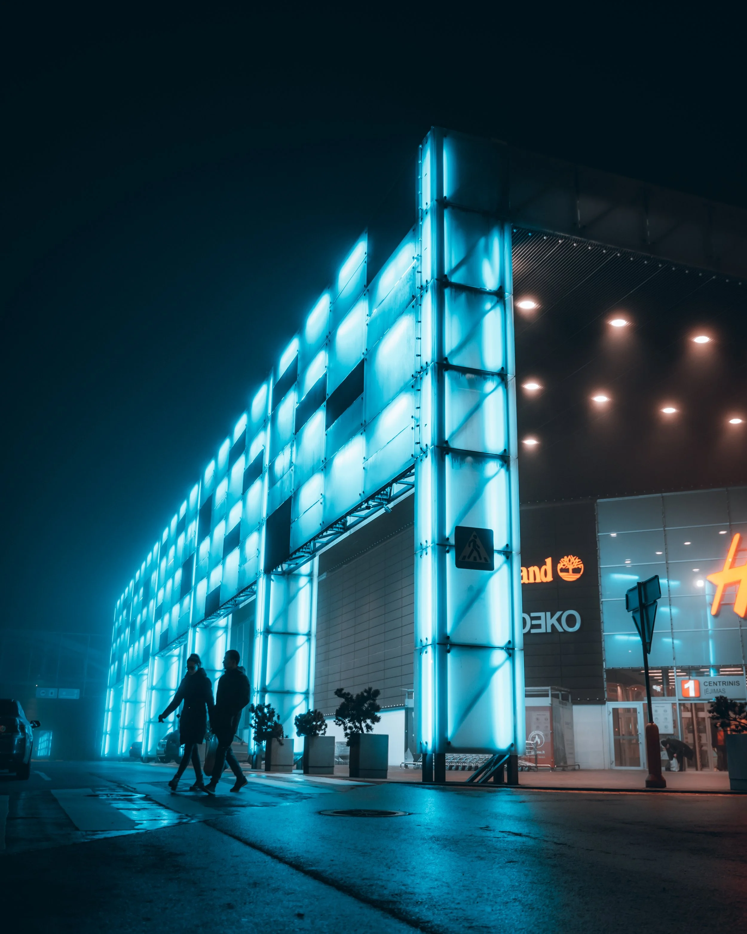 Night view of a shopping mall with bright blue neon lights on the facade, two people walking across the parking lot, and store signs visible in the background.