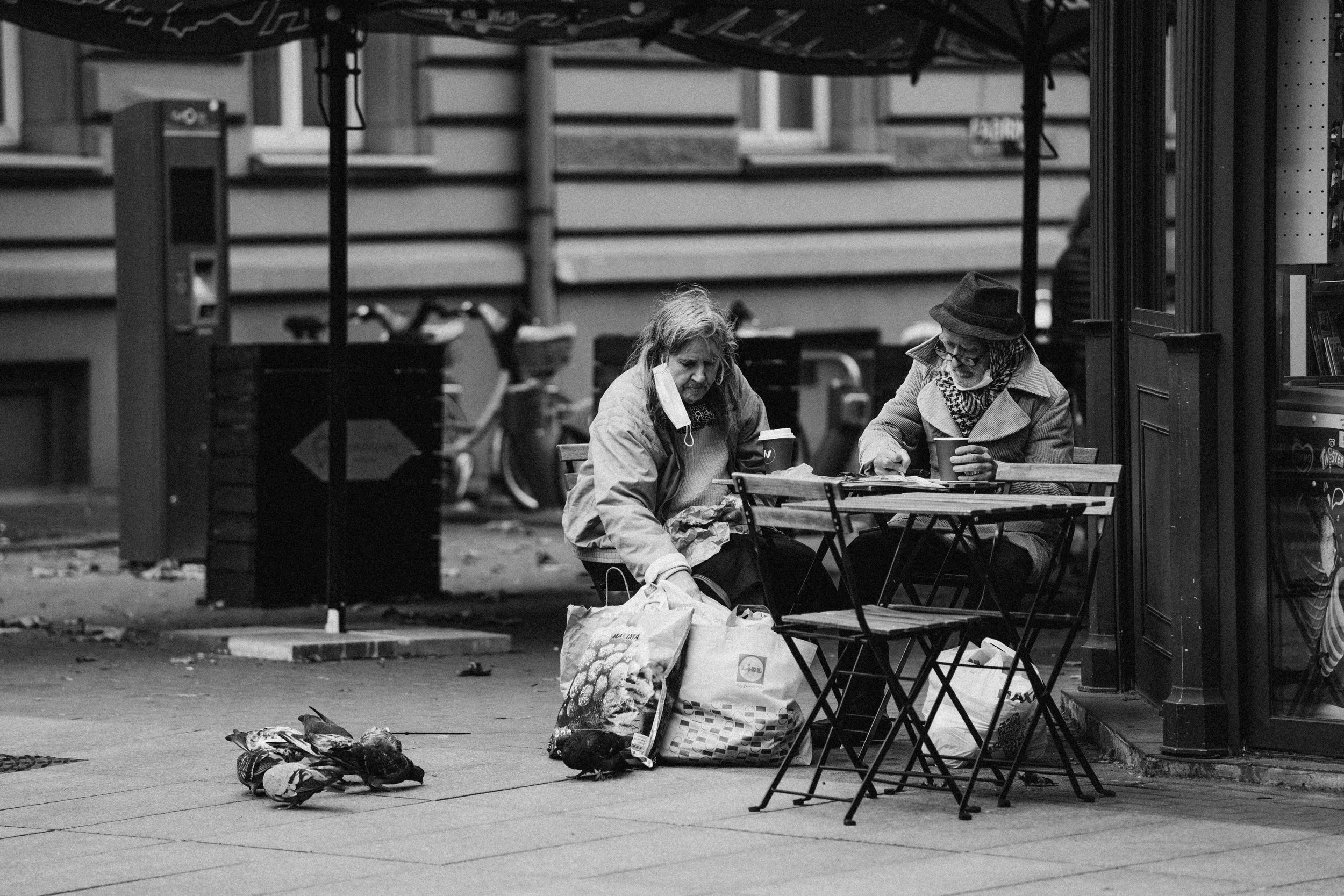 Two homeless individuals, one wearing a coat and the other a hat, sitting at a small table on a city sidewalk, surrounded by bags and pigeons, both holding disposable cups.