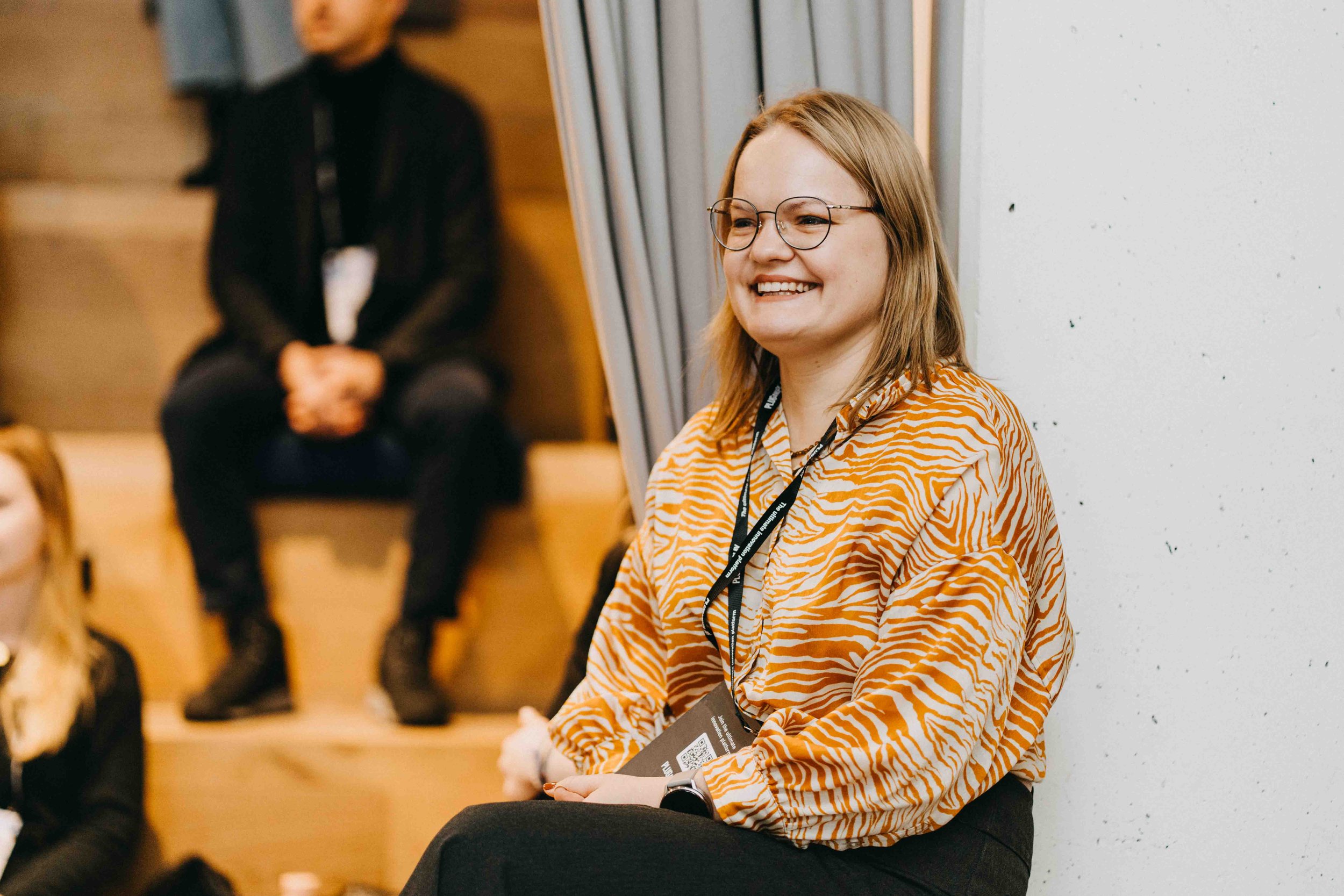 A young woman with shoulder-length blonde hair, glasses, and a bright smile, sitting against a white wall at what appears to be a professional conference or event, wearing a tiger-striped orange and white blouse and a lanyard with an identification b