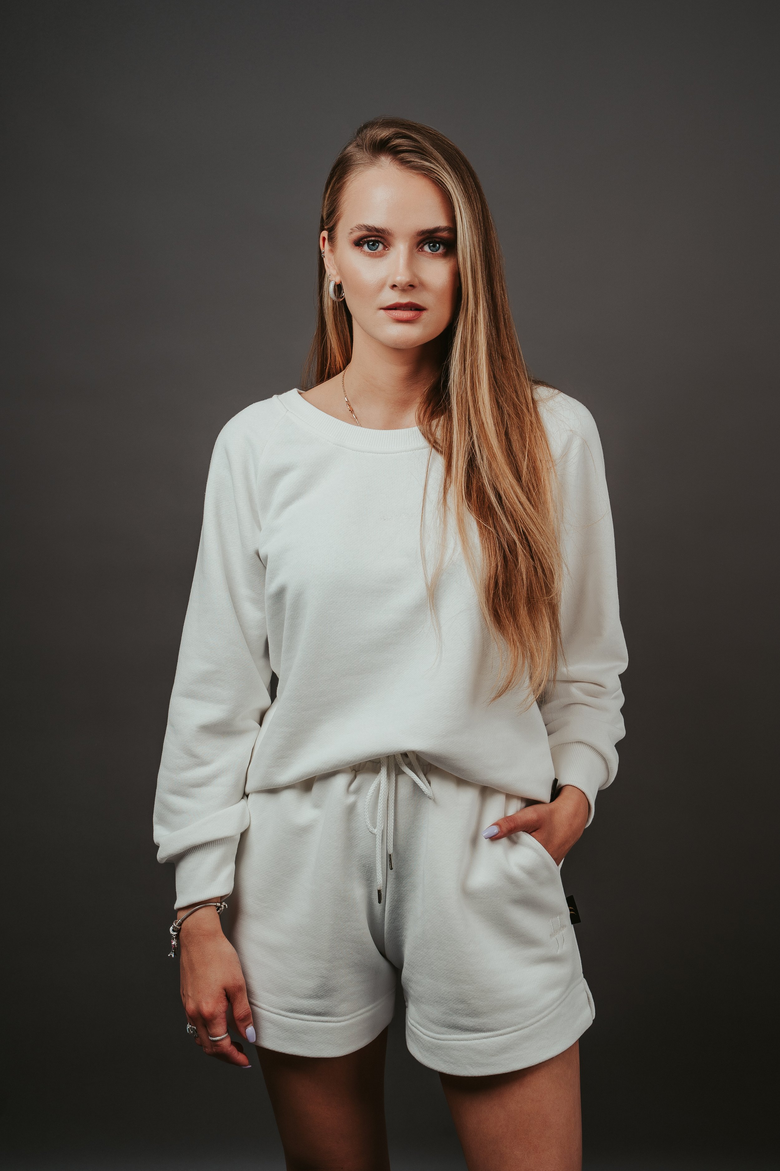 A young woman with long, wavy blonde hair, blue eyes, and makeup, wearing a white sweatshirt and matching shorts, standing against a dark gray background.