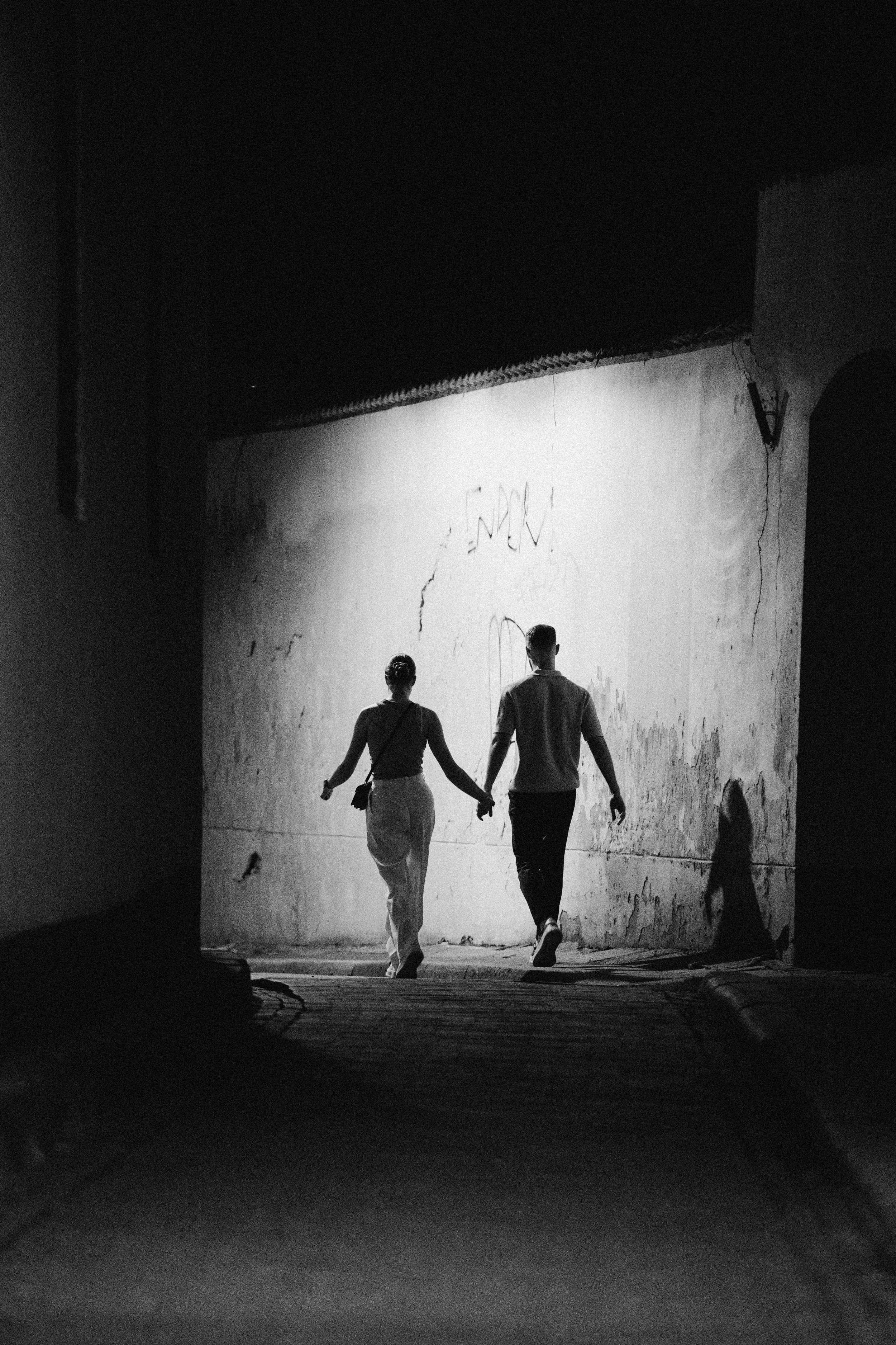 A black and white photo of a couple holding hands while walking in a dark tunnel or alley at night, illuminated by a light source behind them.