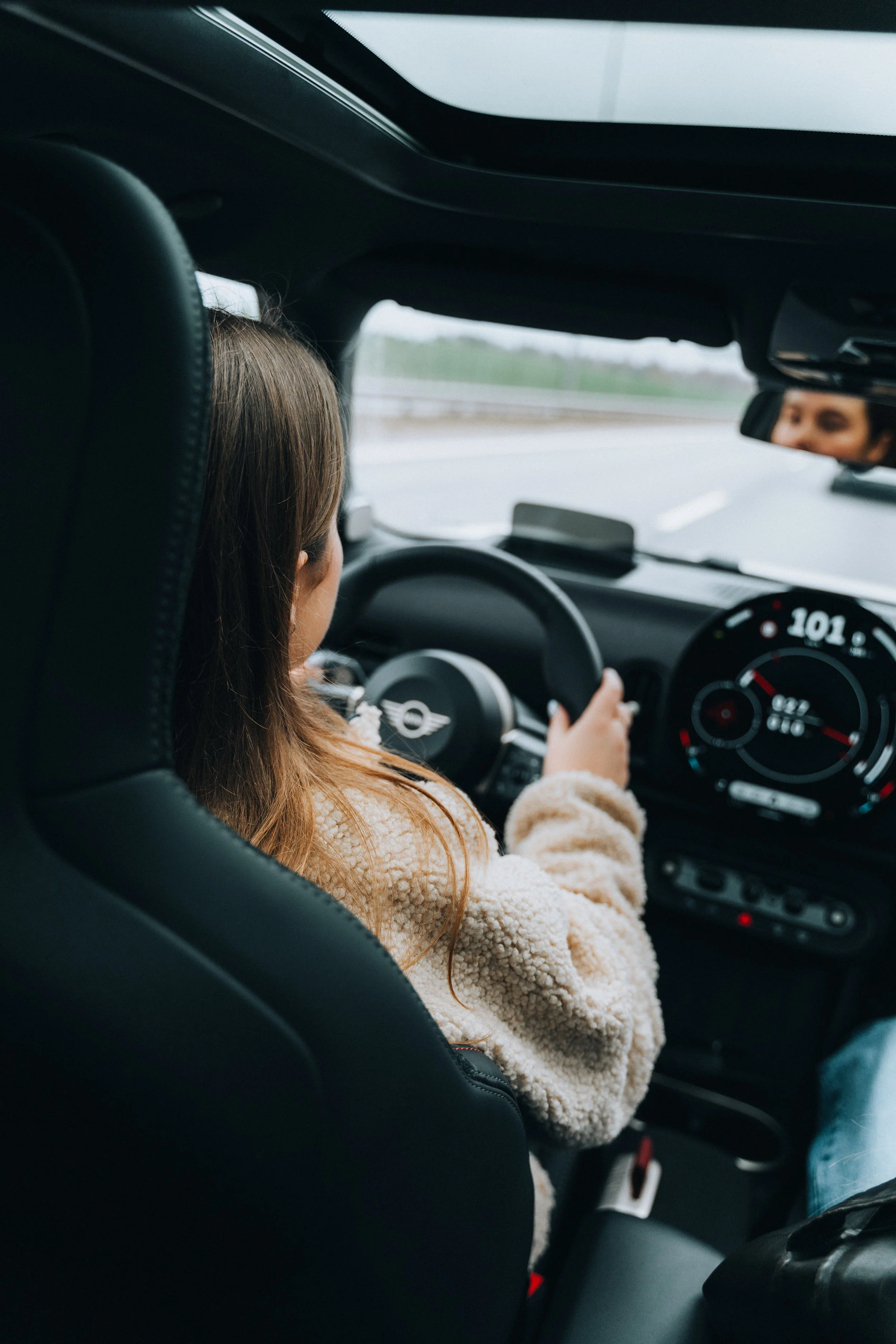 A young girl driving a car with a Mini Cooper logo on the steering wheel on a highway.