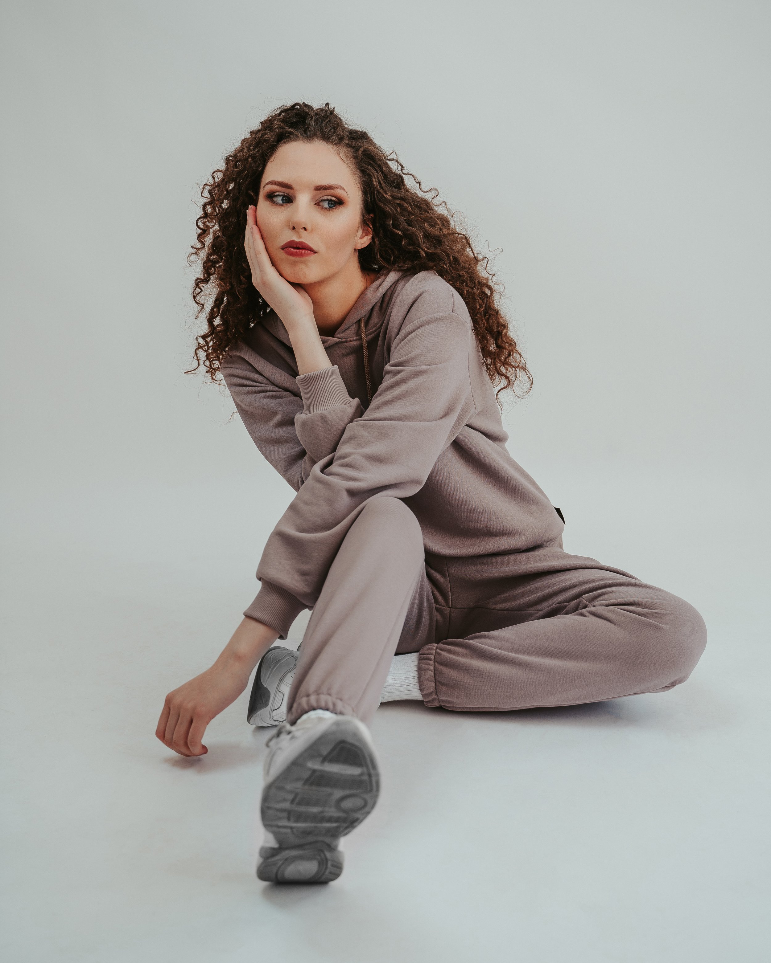 Young woman with curly hair sitting on the floor in a beige tracksuit, resting her head on her hand, looking contemplative.
