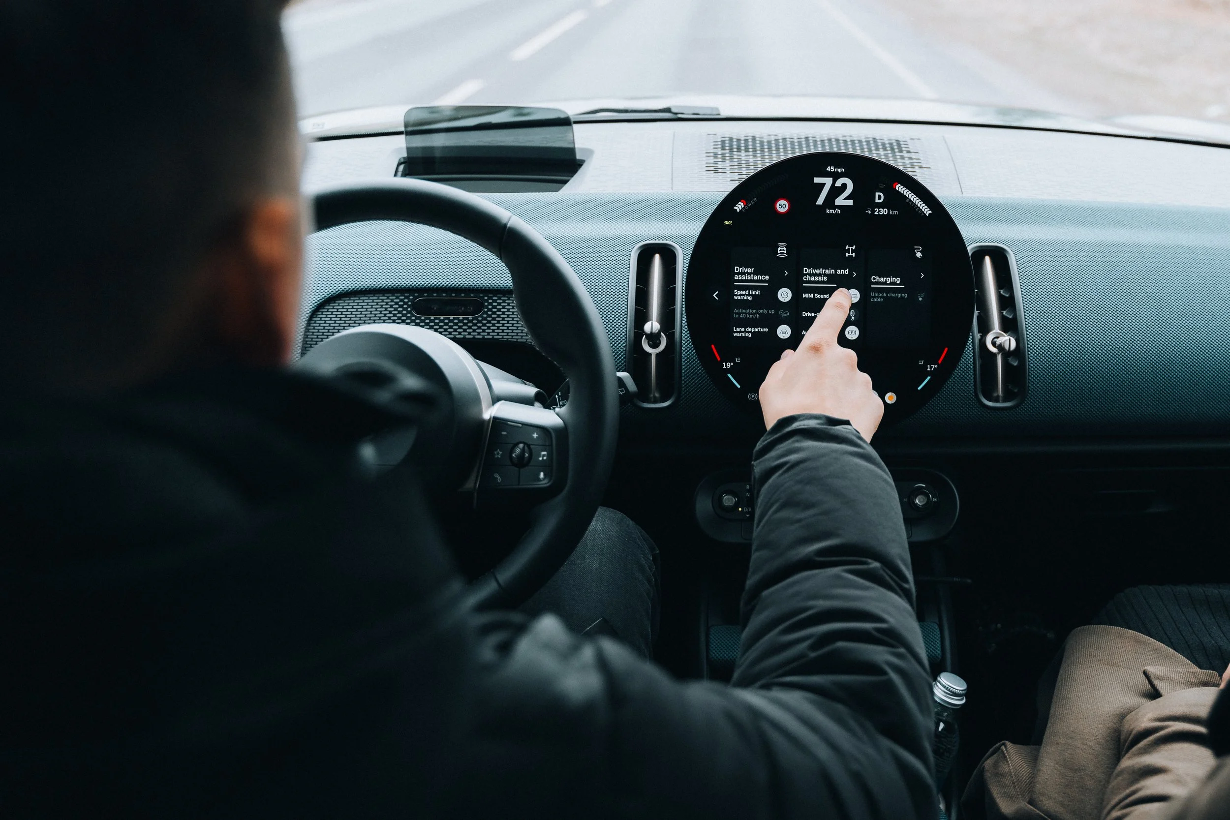 Person in the driver's seat of a vehicle interacting with a digital display on the dashboard, showing driving information and settings.