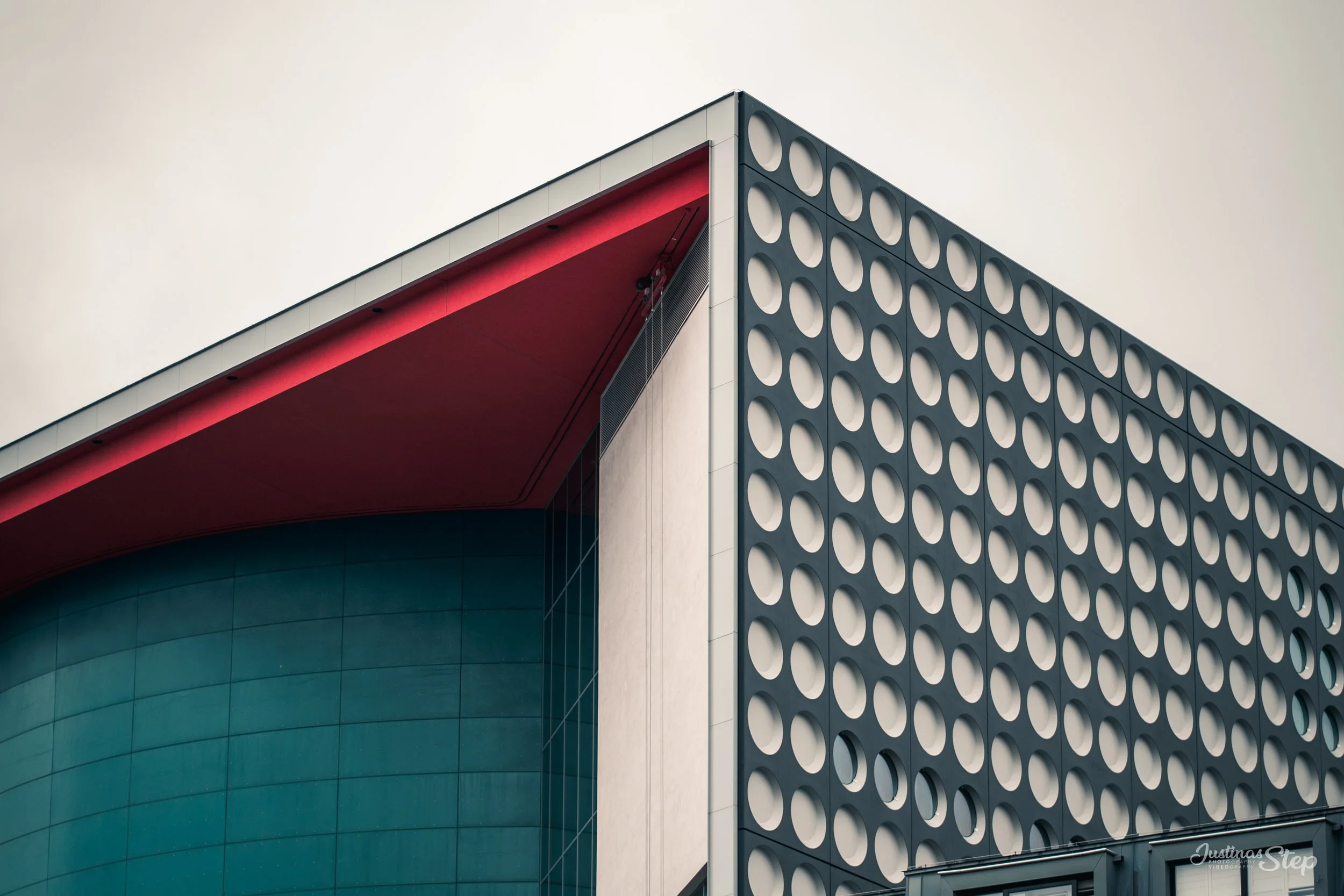 Close-up of a modern building's corner showing a section with circular cut-out patterns, a curved teal wall, and a red underside of the roof.