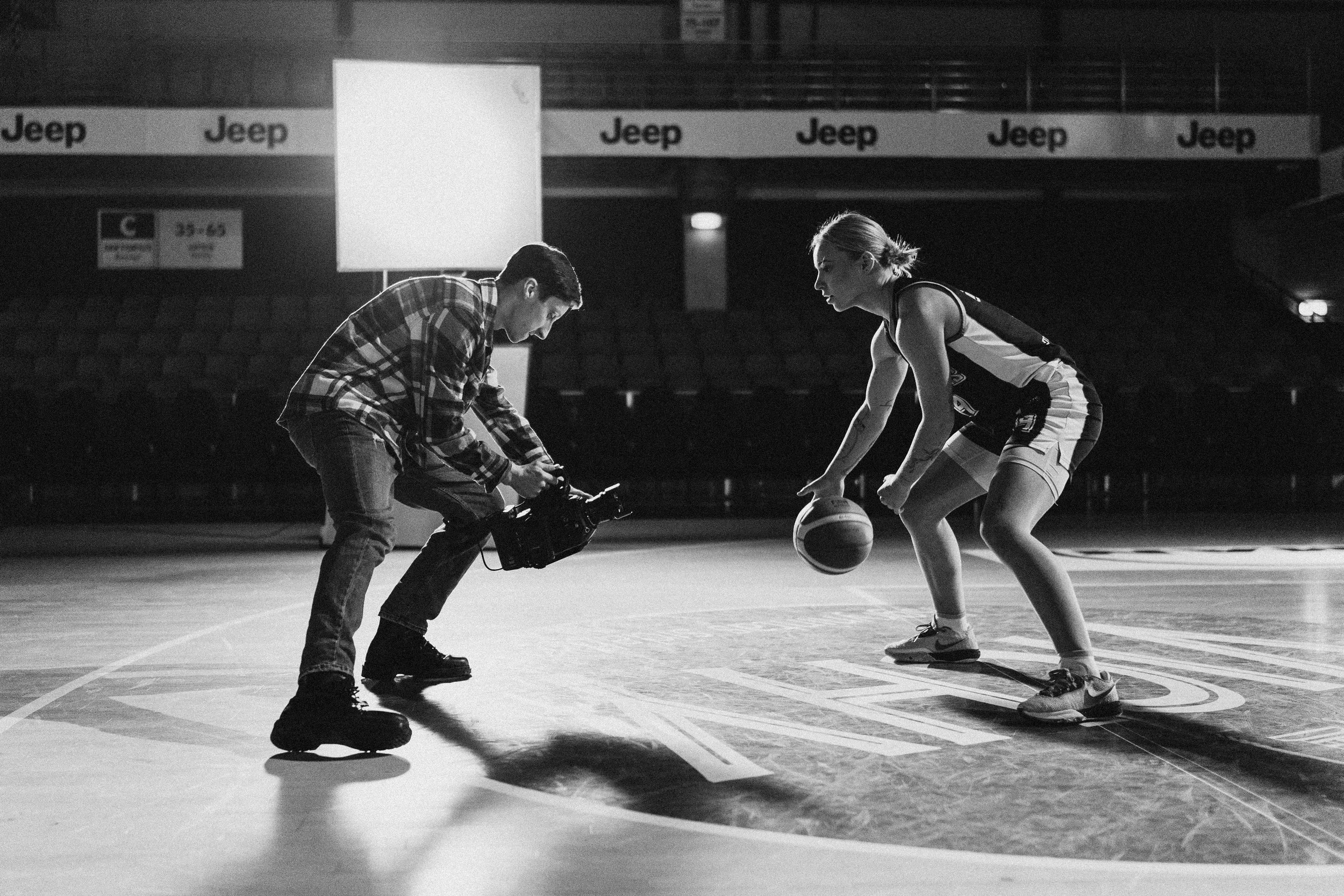 A woman in sportswear practicing basketball on a court with a camera operator filming her, in an indoor stadium with 'Jeep' advertisements.