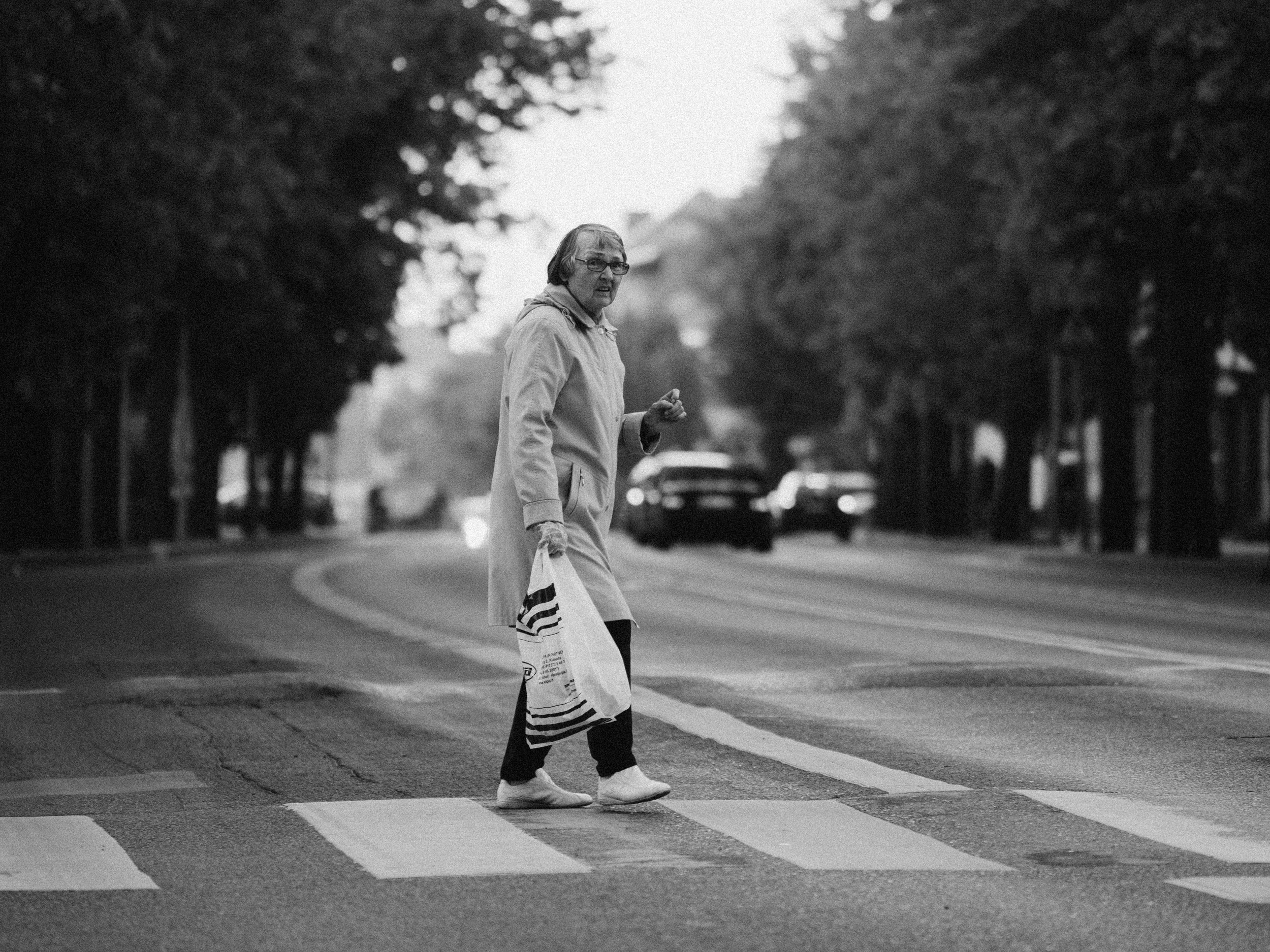 An elderly woman with glasses, wearing a raincoat, walking across a zebra crossing while holding a shopping bag.