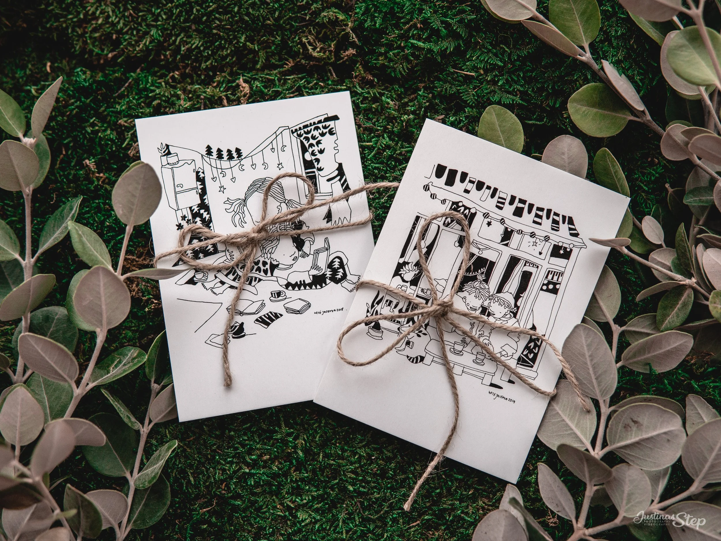 Black and white illustrated holiday greeting cards tied with twine, lying on green moss surrounded by eucalyptus branches.