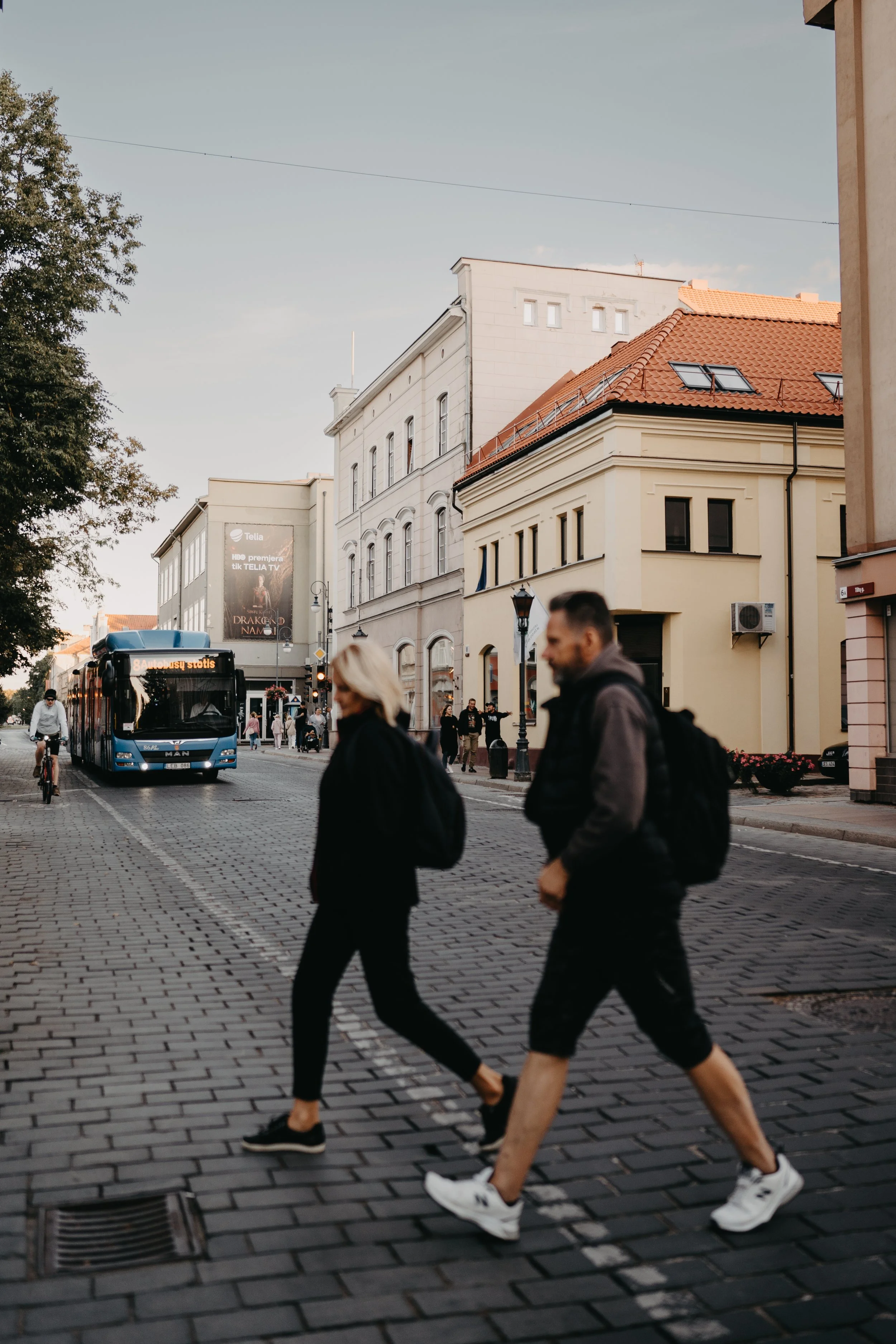 Street scene with pedestrians crossing, a blue city bus, cyclists, and historic buildings during early evening in a European city.