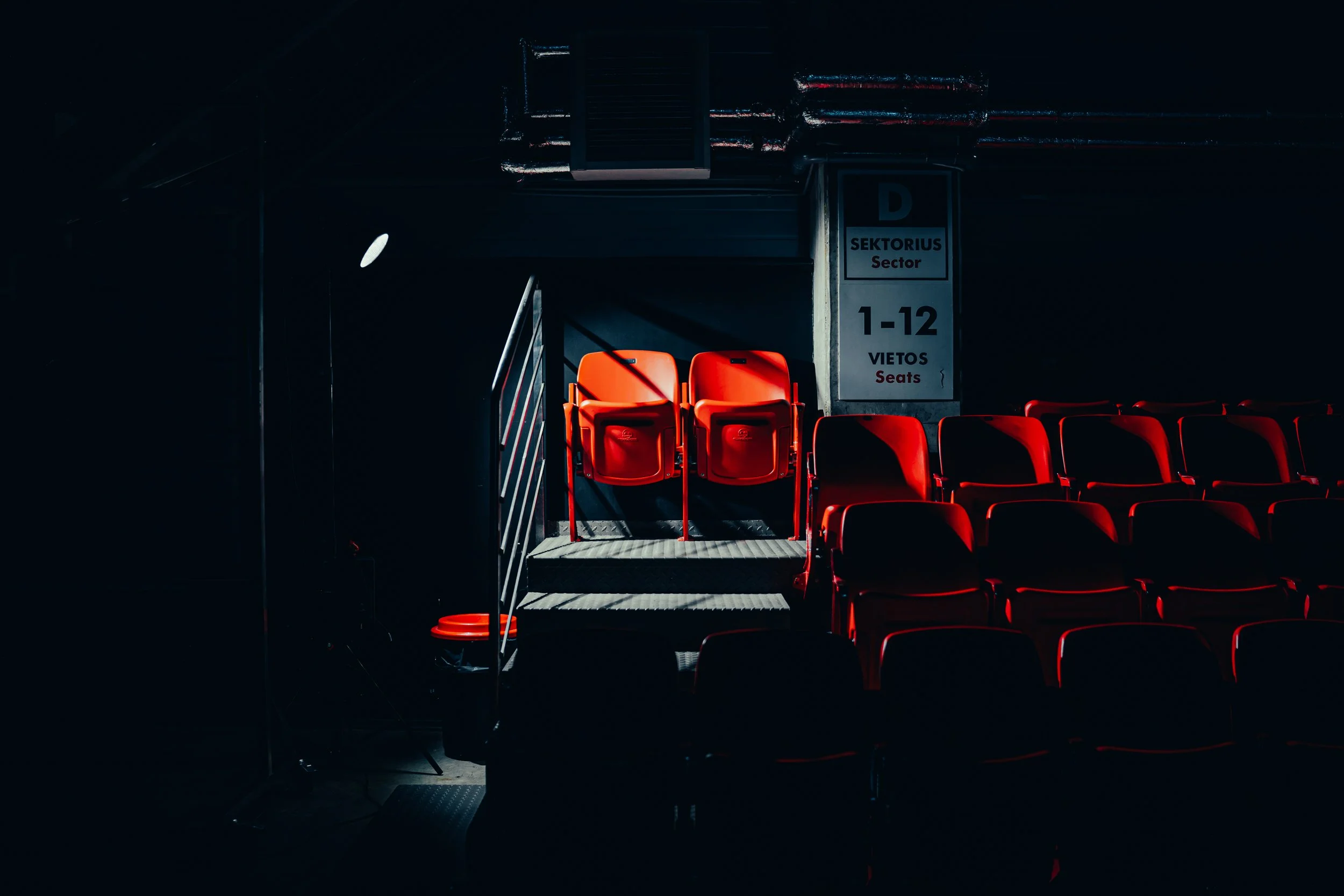 Red chairs in a dark theater or auditorium, with some seats illuminated by light, and a sign indicating sector D, seats 1-12.