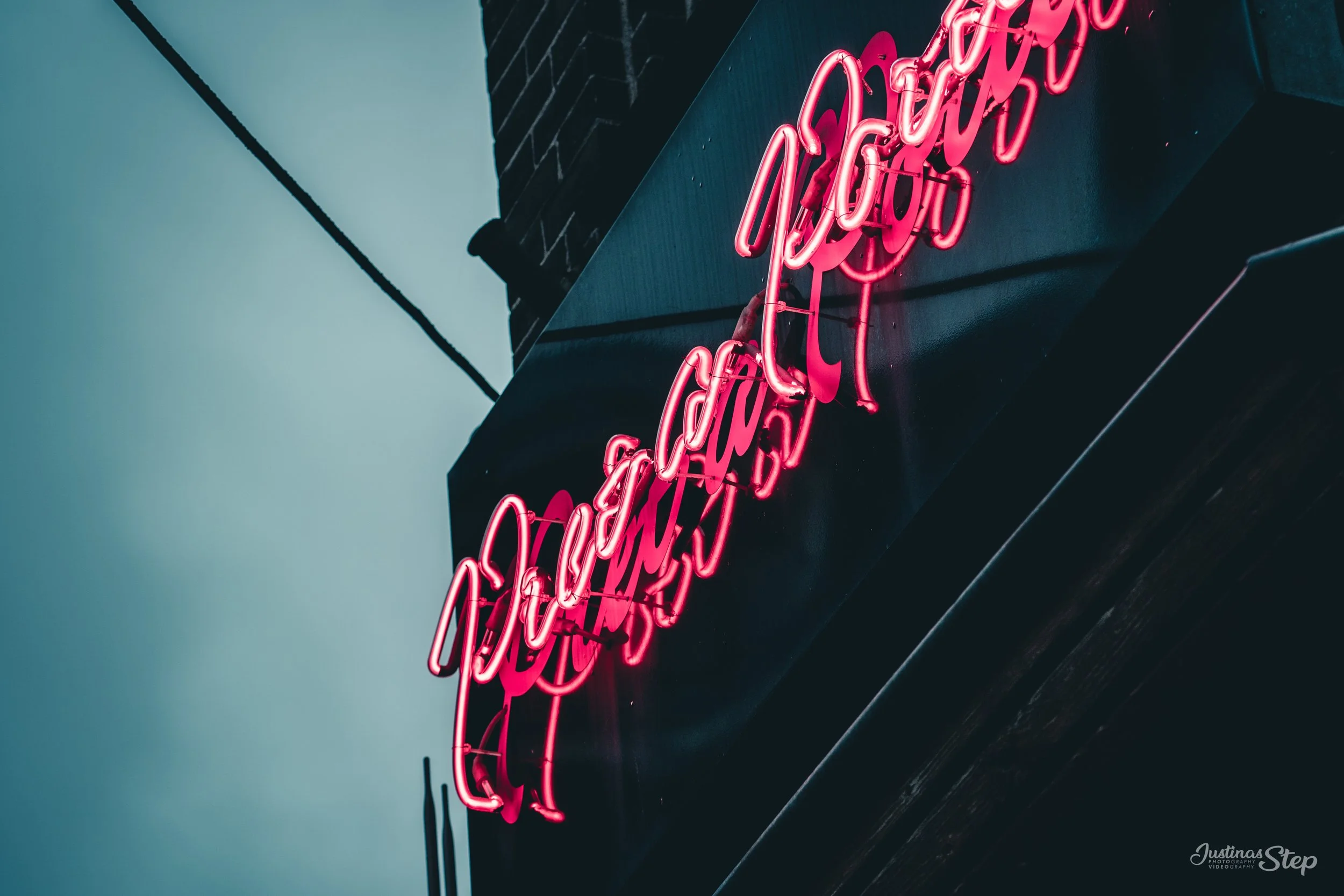Pink neon sign on a black building with a brick accent, partially visible, against a gray sky.
