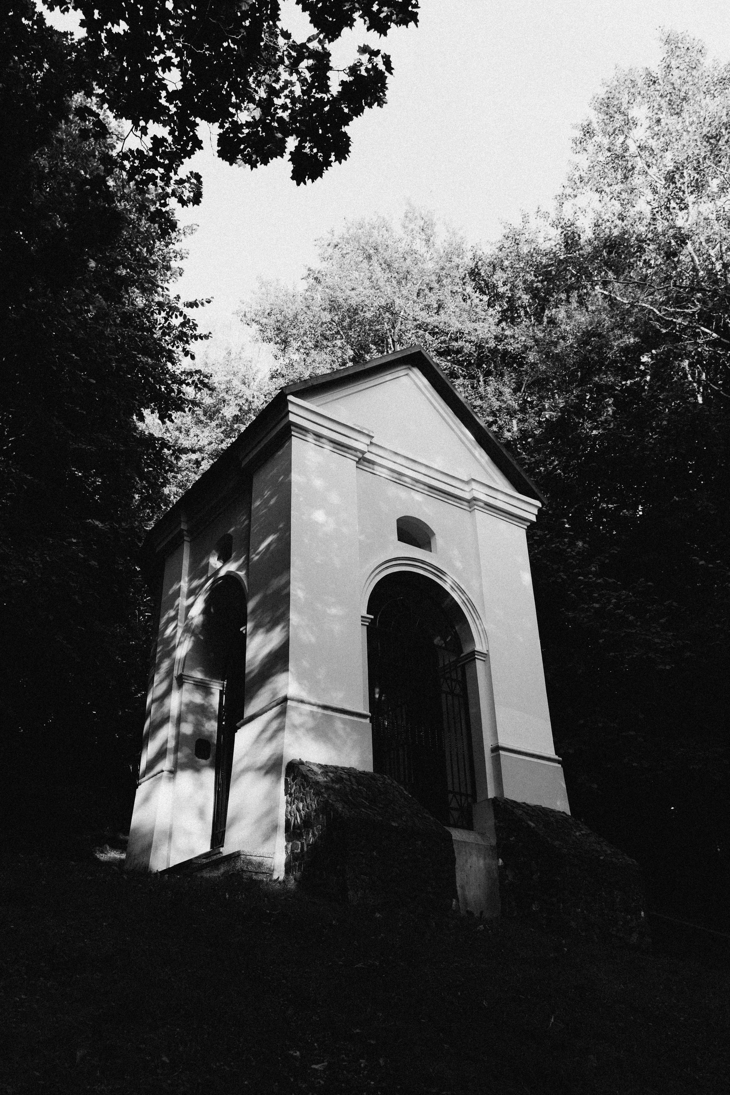 A white architectural structure with arches, set against a backdrop of trees and the sky, with shadow patterns on its surface.